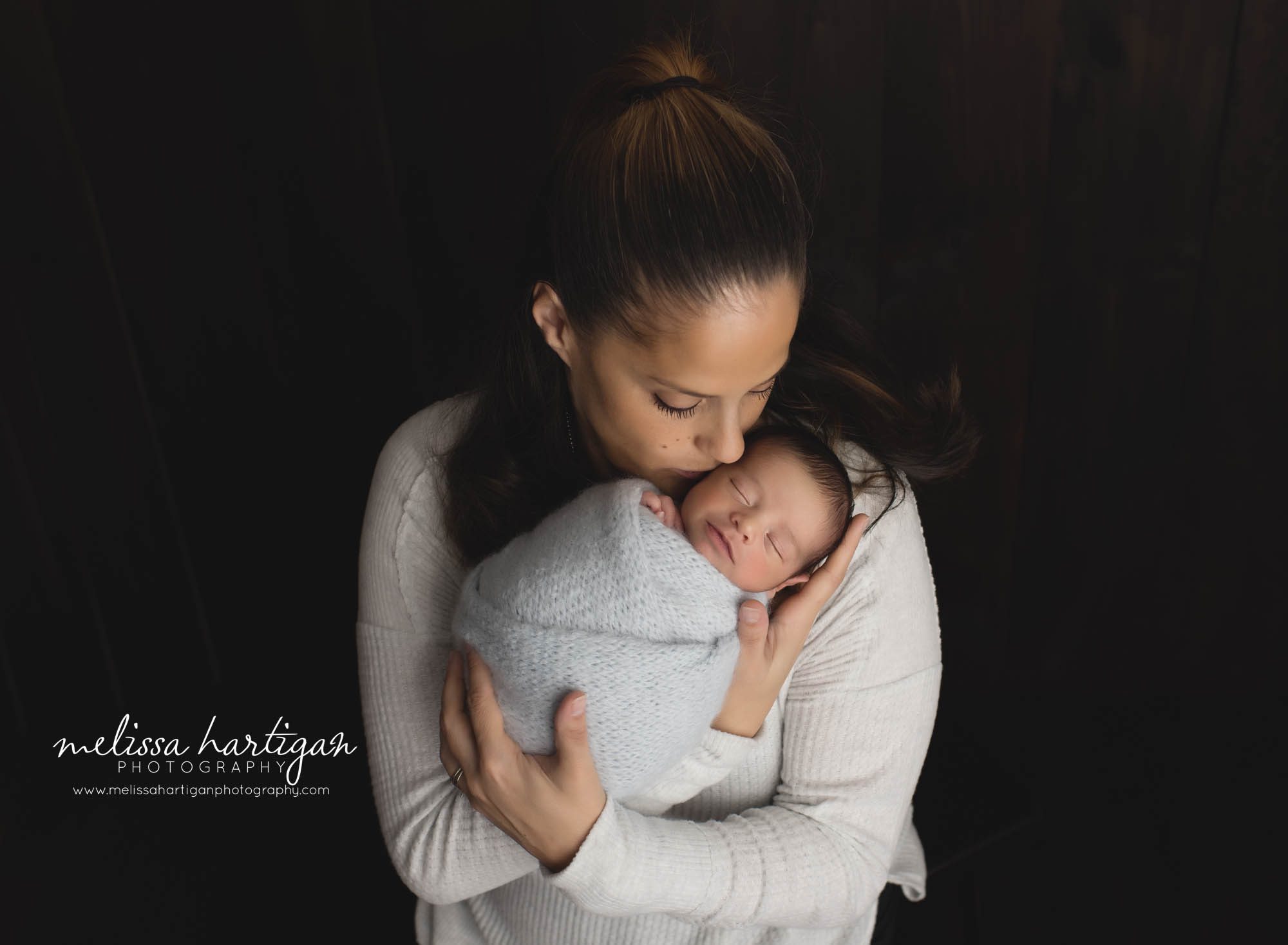 mom holding newborn son wrapped in light blue knitted wrap kissing him on the cheek CT newborn Photography