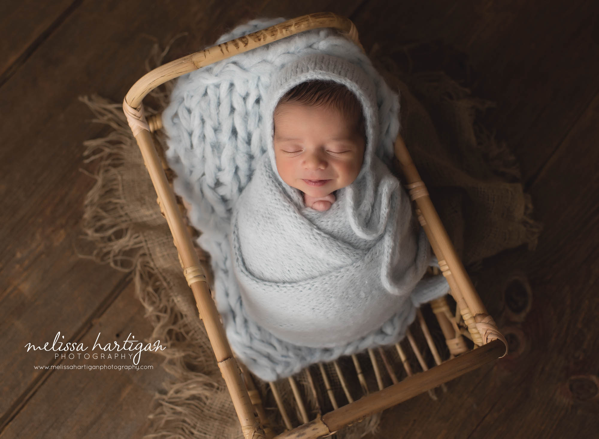 newborn baby boy wrapped in light blue knitted wrap with matching bonnet baby in wooden wicker basket smiling baby newington ct baby photographer