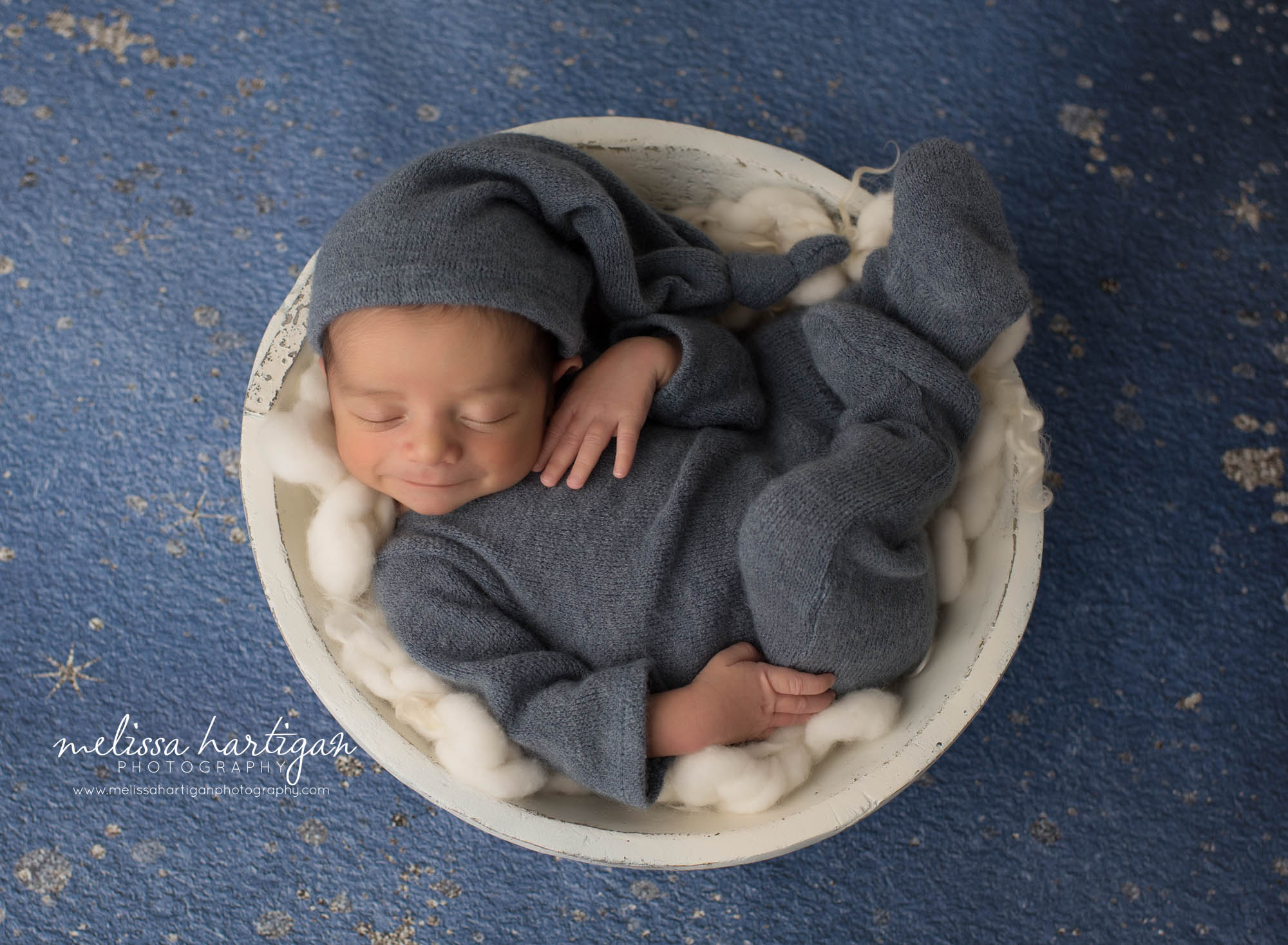newborn baby boy wearing footed sleeper with matching sleepy cap posed in cream wooden bowl with cream layers and smiling newington newborn photography
