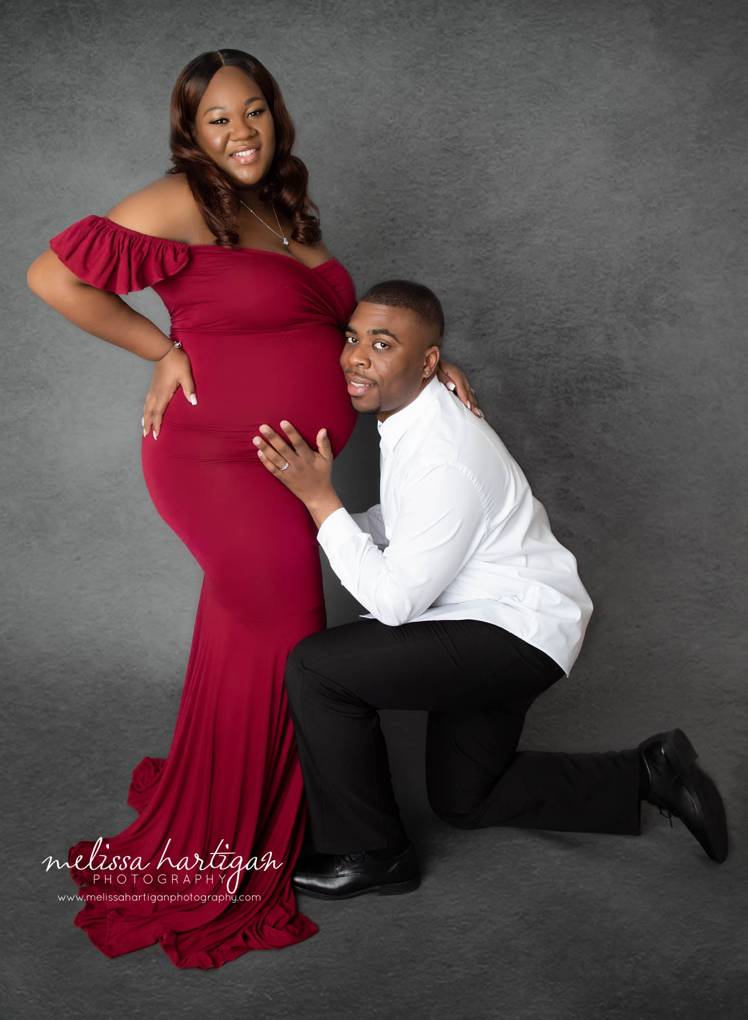 dad kneeling down in studio with head to mom to be's pregnant baby belly mom wearing red maternity dress MA materntiy photography CT