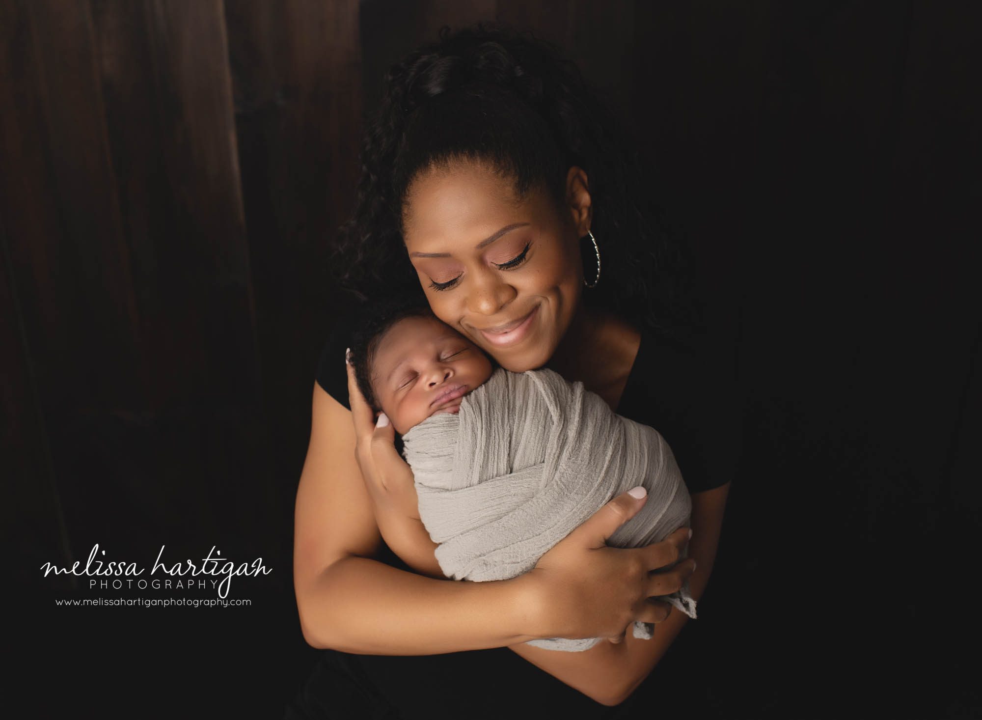 mom smiling holding newborn son wrapped in gray wrap
