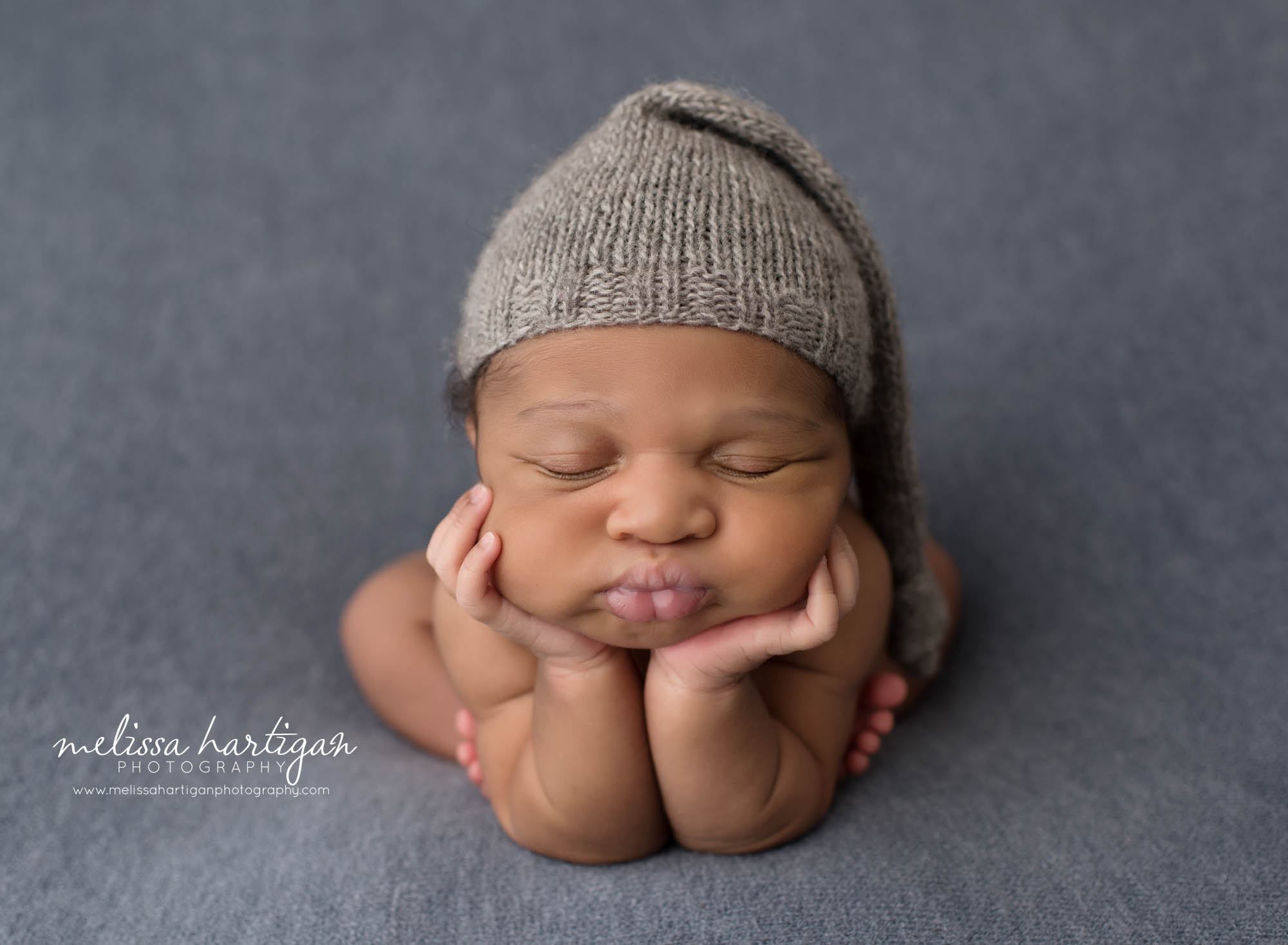 newborn baby boy posed froggy pose with puckered lips on blue backdrop wearing gray knitted sleepy cap
