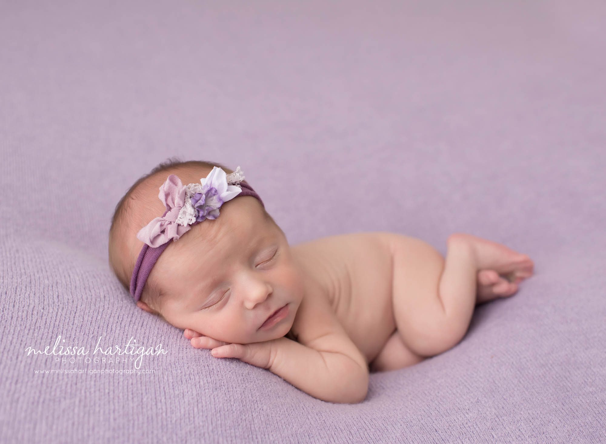baby girl posed on purple backdrop with hand under chin wearing flower bow headband
