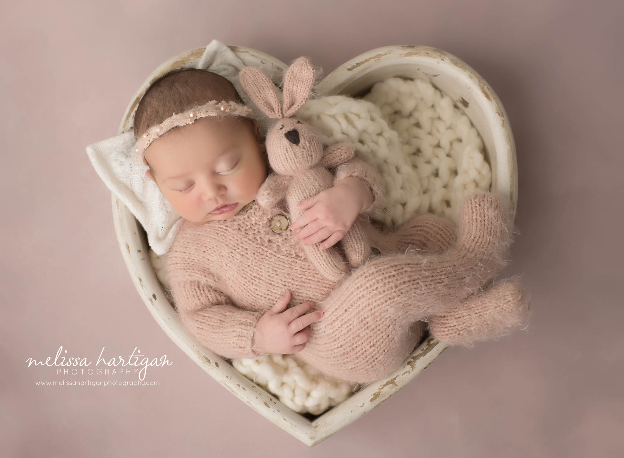 newborn baby girl posed in cream wooden heart shaped prop wearing pink knitted footed sleeper holding knitted bunny prop