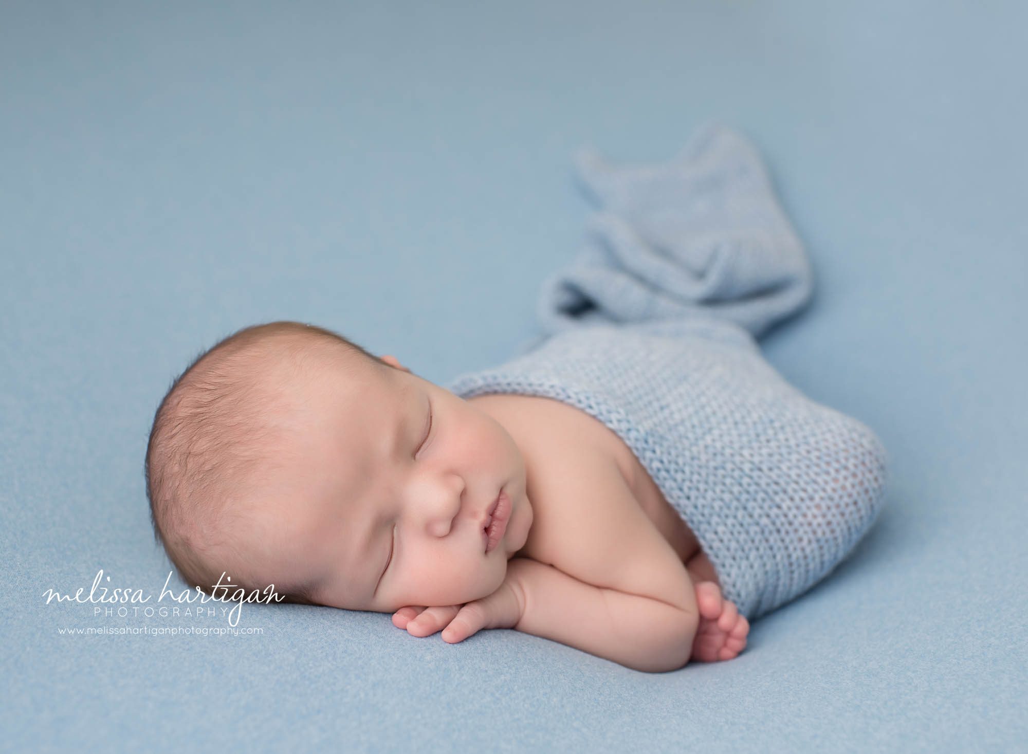 newborn baby boy posed modified taco pose with knitted blue wrap on blue backdrop sleeping baby boy