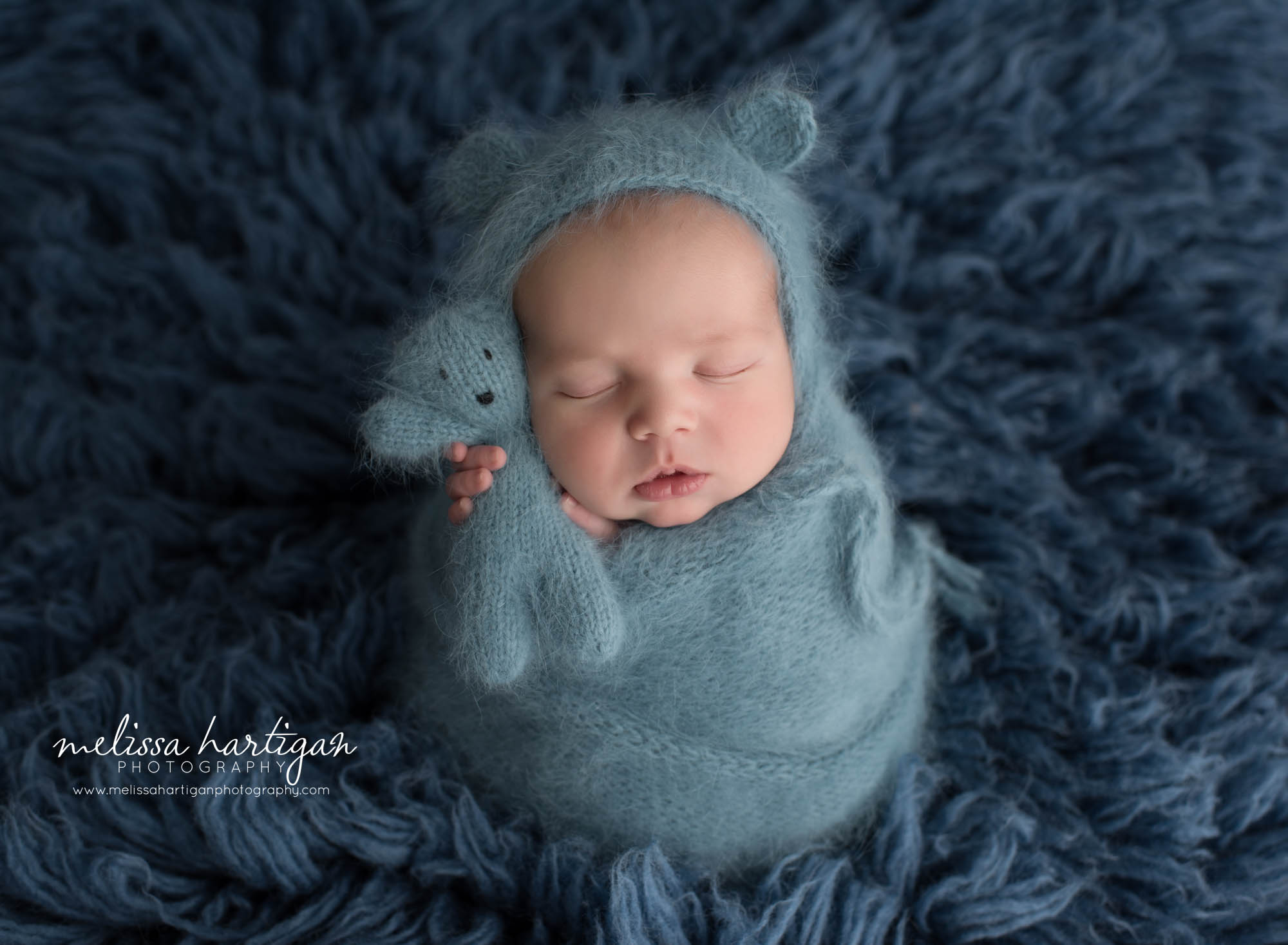 newborn baby boy wrapped in teal blue knitted wrap with matching teddy bear and bear bonnet angora knit pose don dark blue flokati Connecticut newborn photography