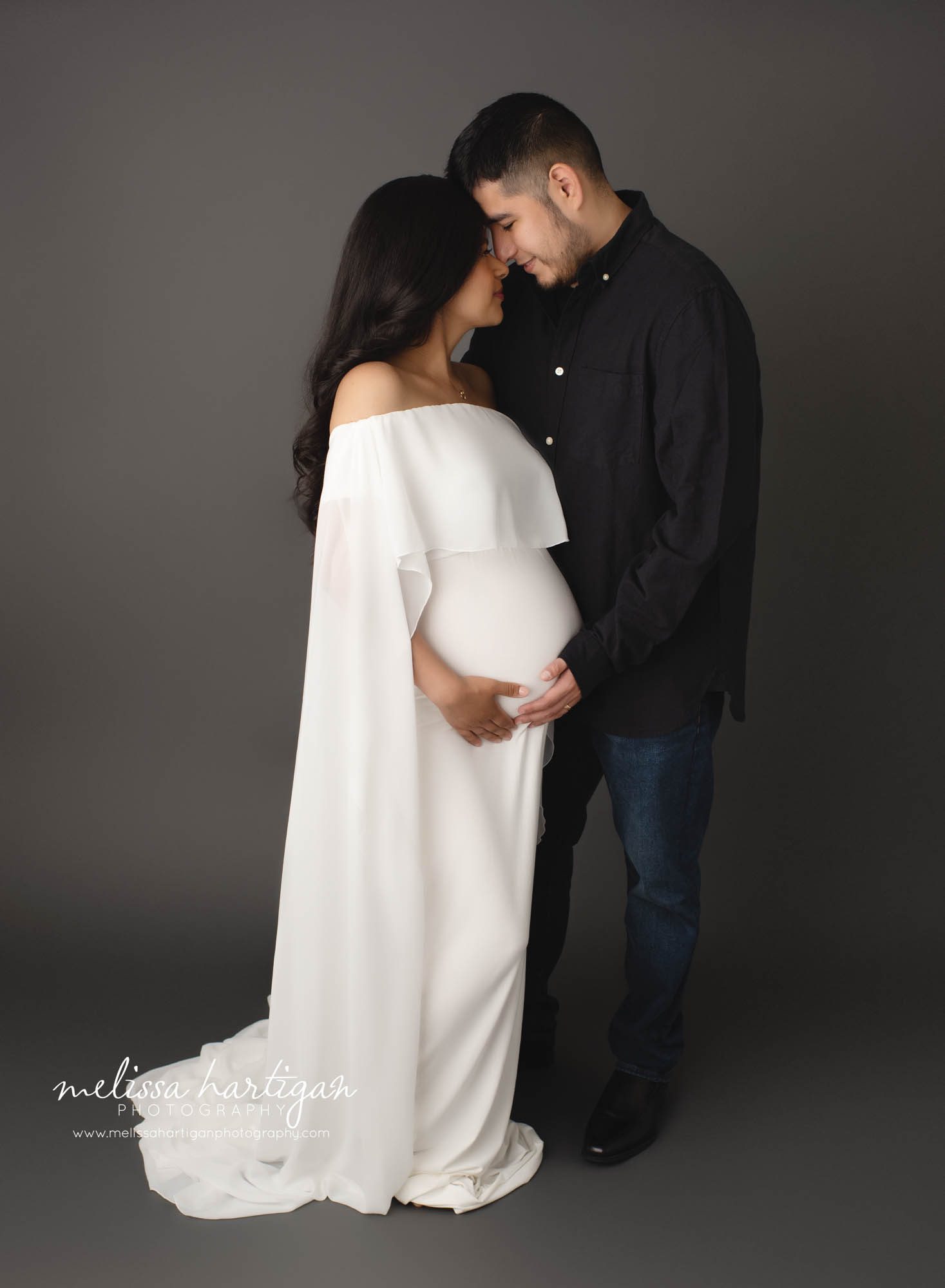 pregnant mom with dad to be standing forehead to forehead holding baby bump in studio pregnany photography session