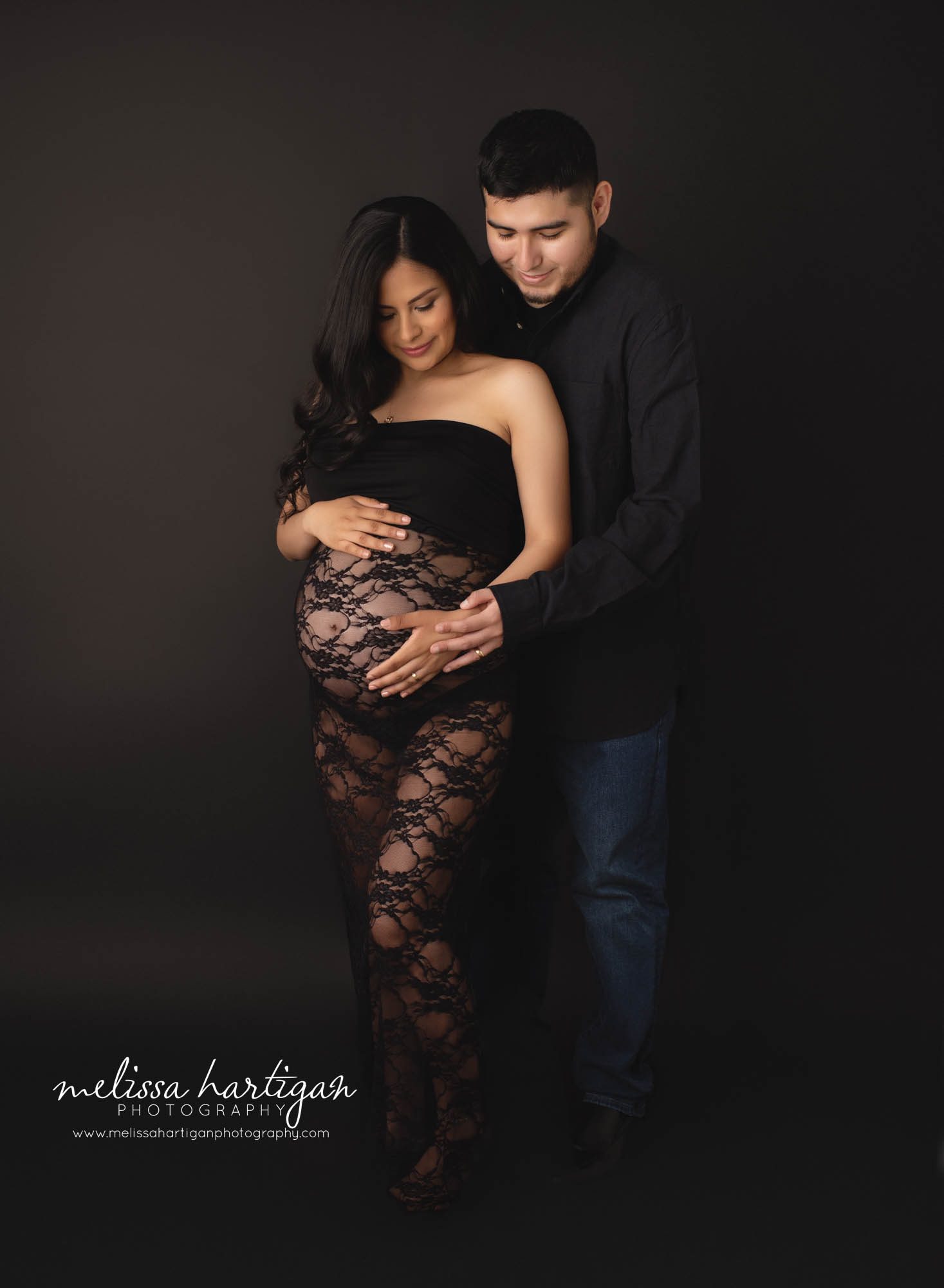 mom and dad to be standing in photography studio holding baby bump looking down happy smiling at pregnant baby belly