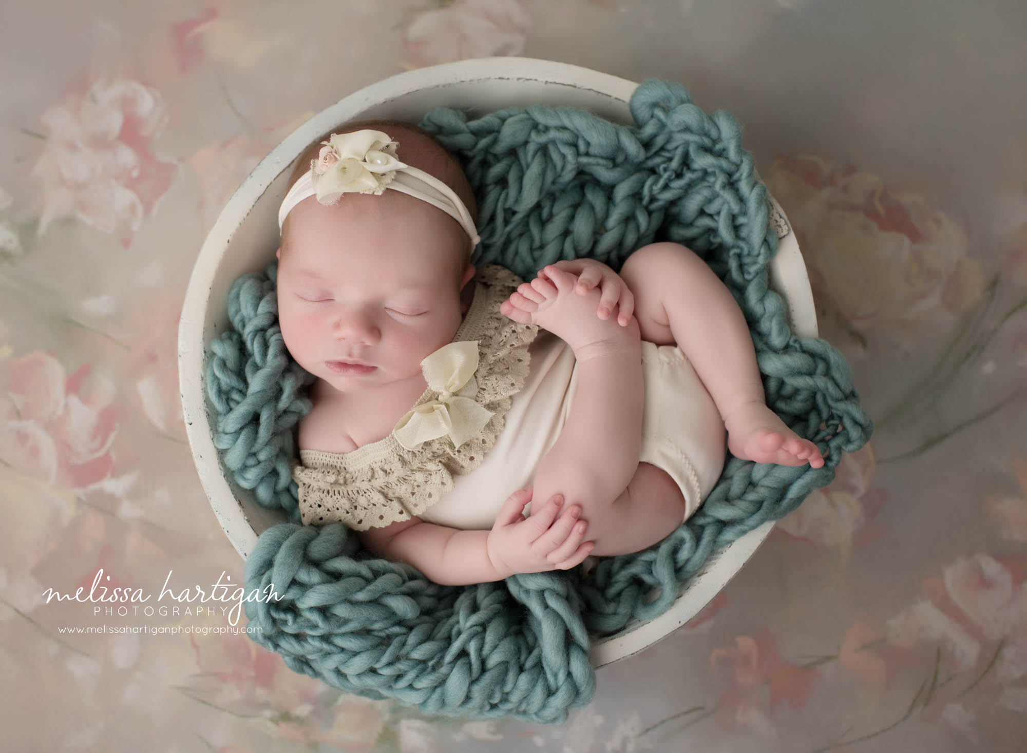 newborn baby girl posed in wooden cream bowl with teal knitted chunky layer wrap wearing cream romper outfit with flower headband