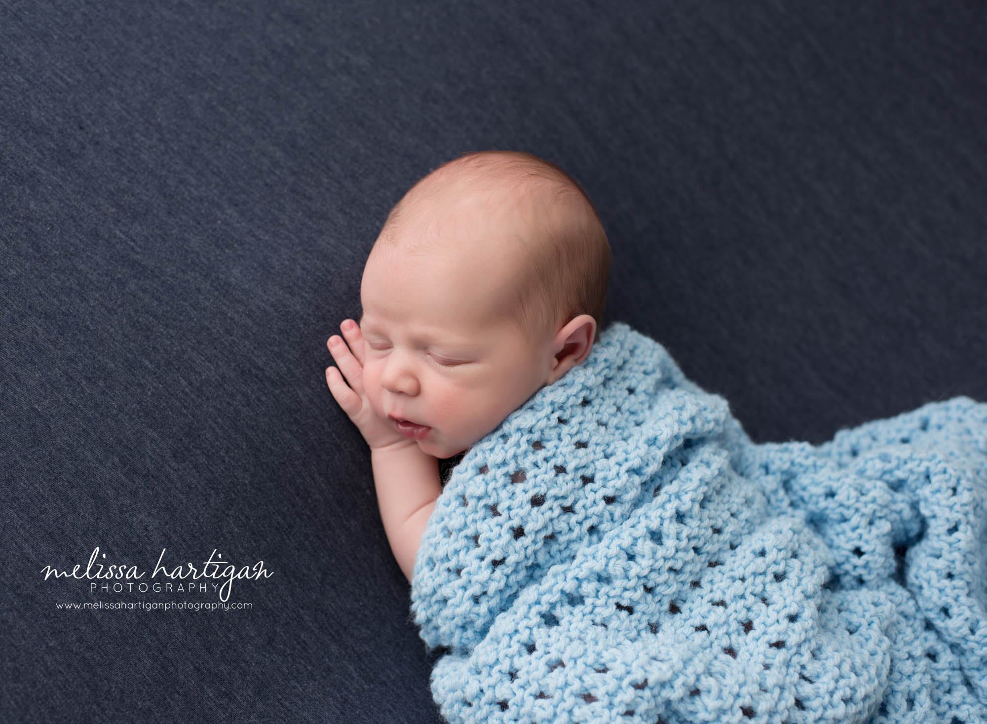 newborn baby boy posedf on blue backdrop with knitted blue baby blanket wrapped around him Newborn Photography Middlesex county