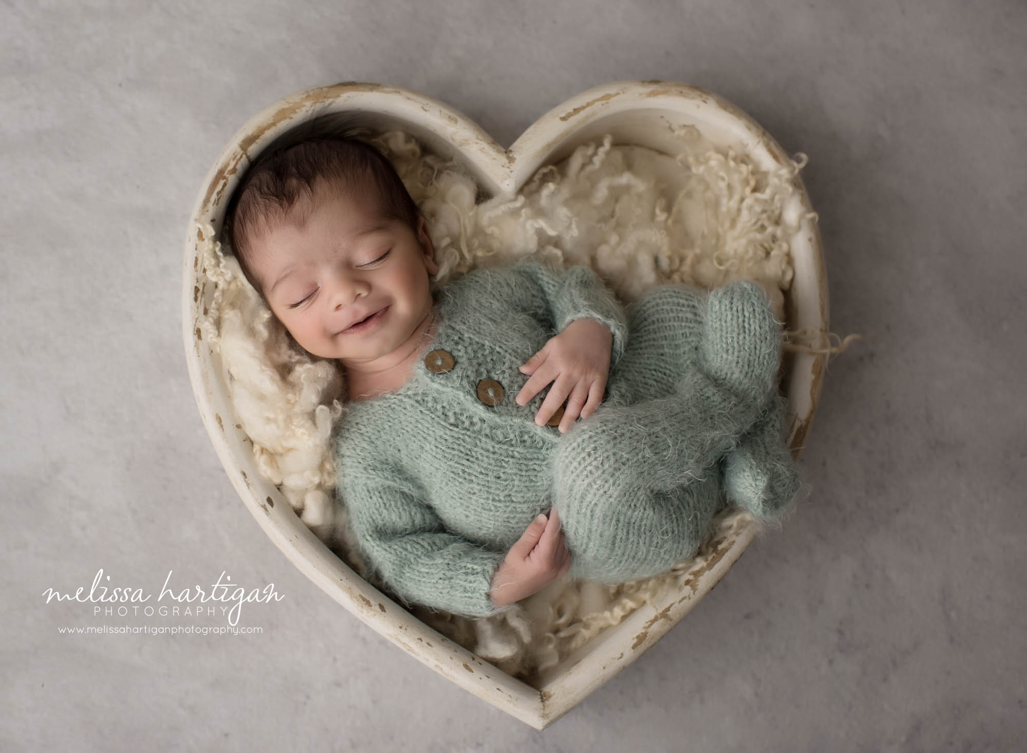 newborn baby boy wearing teal outfit smiling posed in heart shaped wooden bowl prop