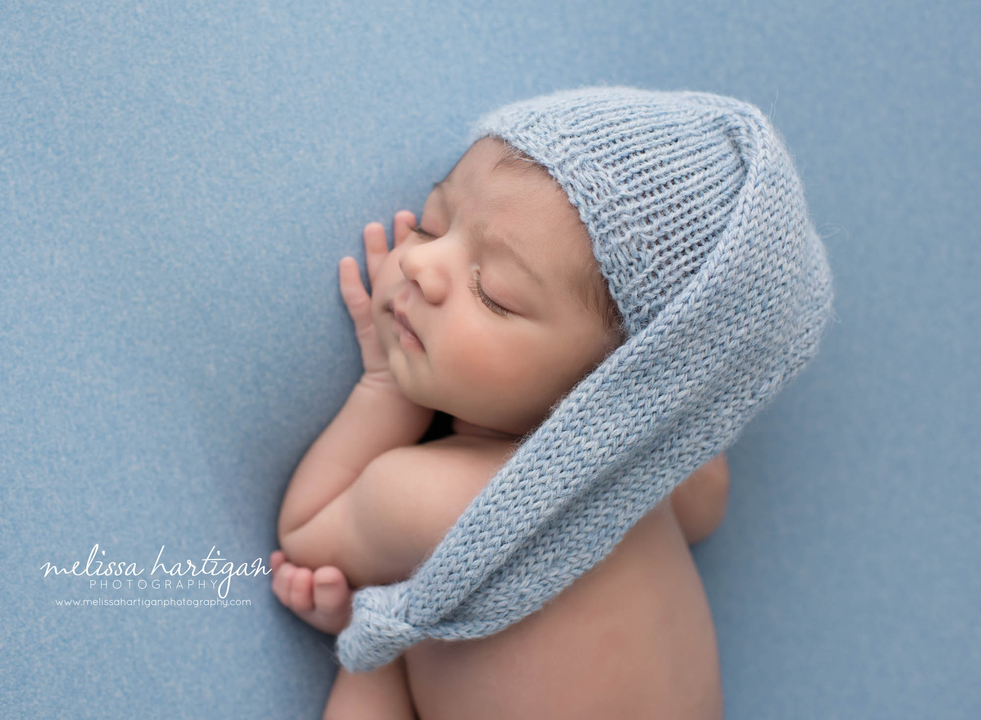 newborn boy posed on blue backdrop wearing knitted blue sleepy cap