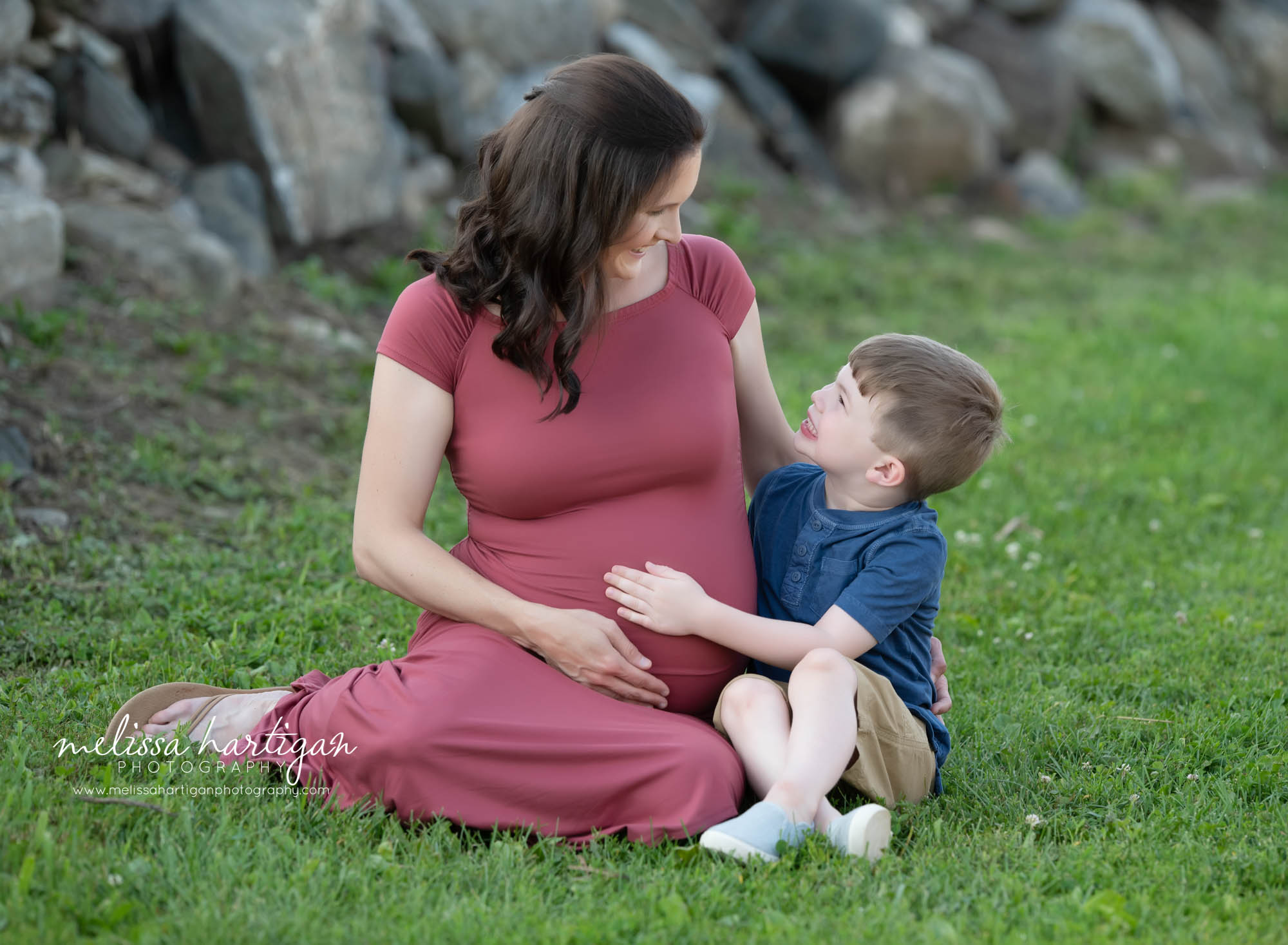 mom sitting on grass with son holding baby belly