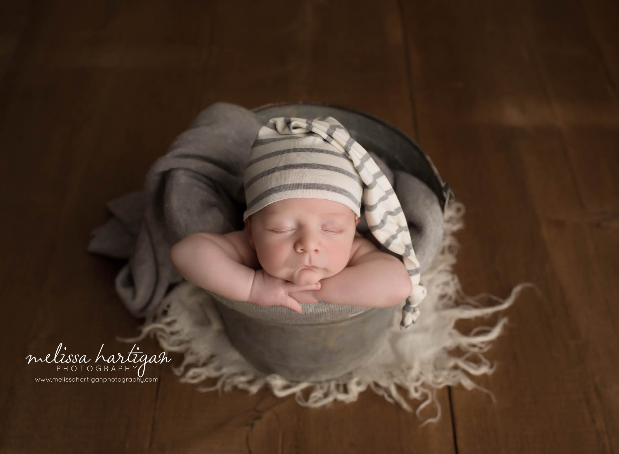 newborn baby boy posed in bucket wearing striped sleepy cap with gray layer wrap