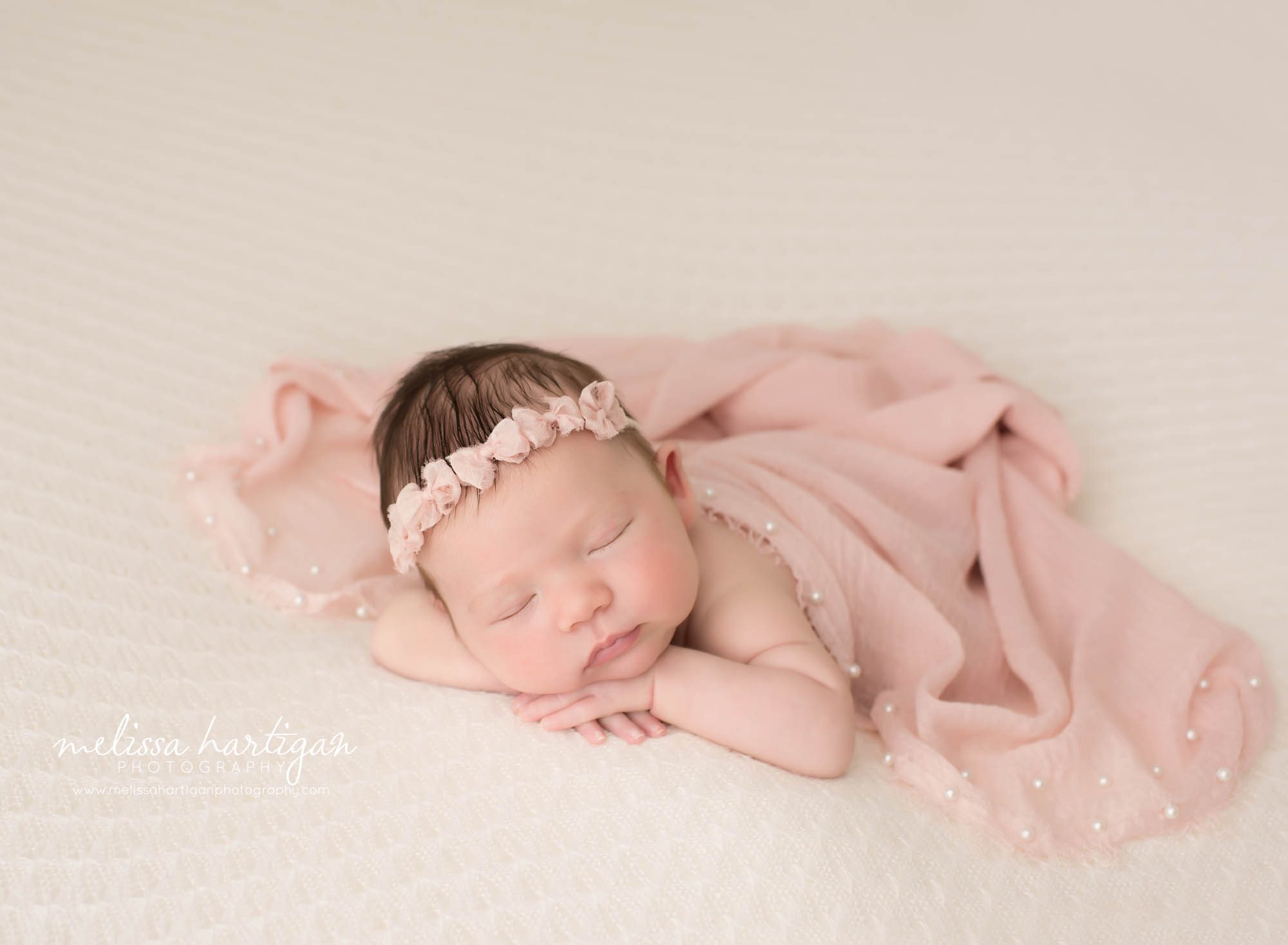 newborn baby girl posed on tummy with pink wrap draped over her back wearing bow headband