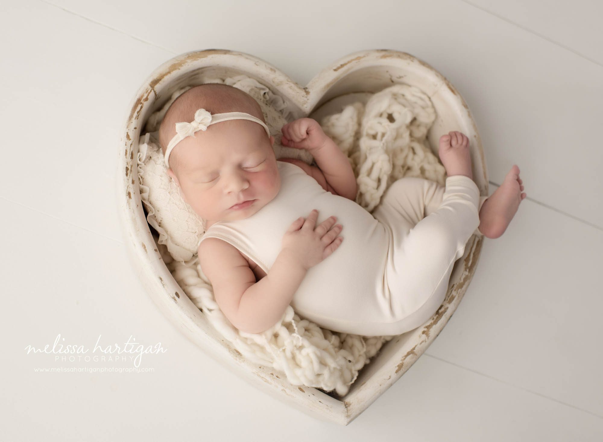 newborn baby girl posed in heart prop wearing white cream outfit with bow headband