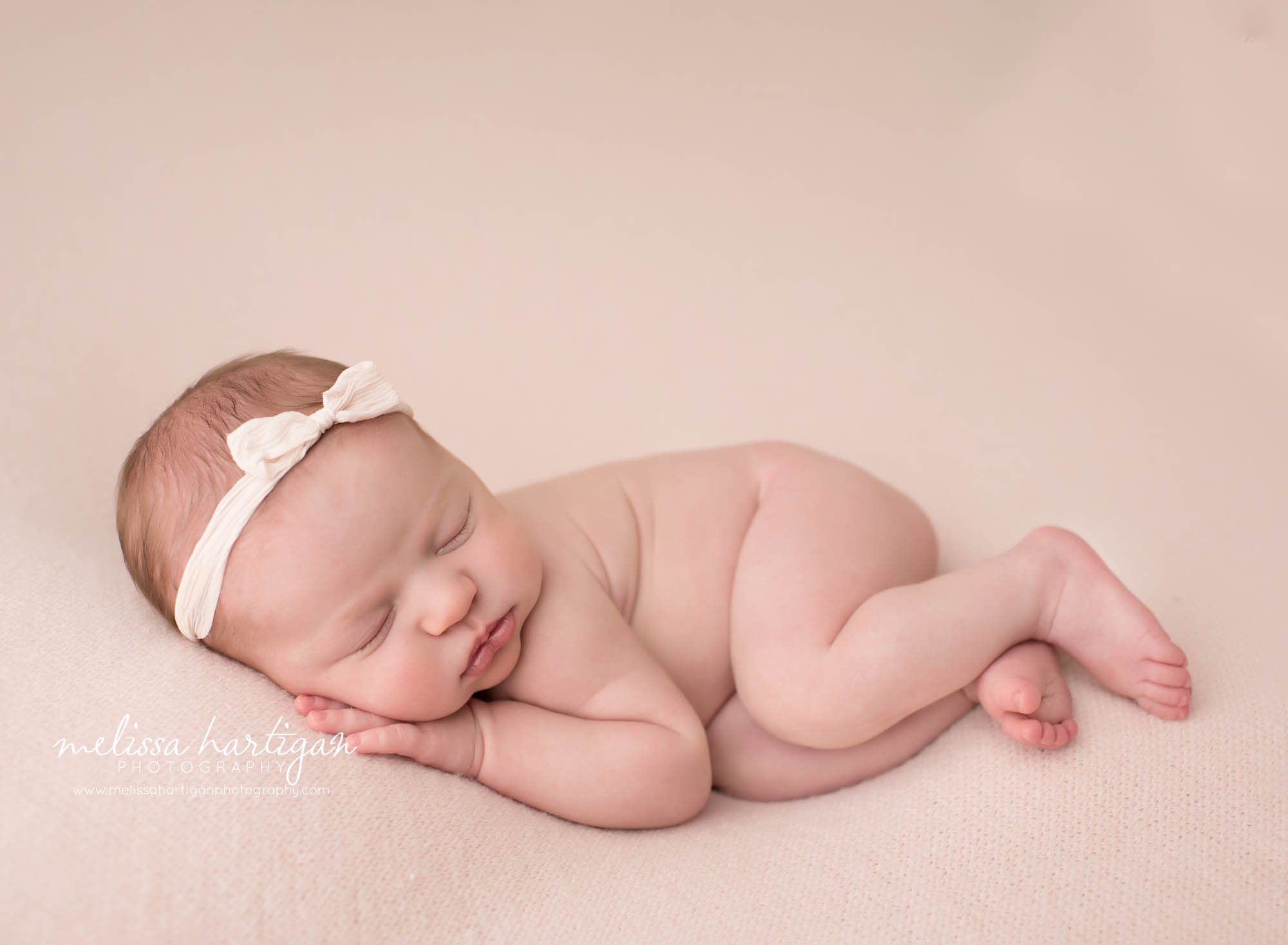 newborn baby girl posed on pink backdrop on tummy side with hand under cheek wearing pink bow headband