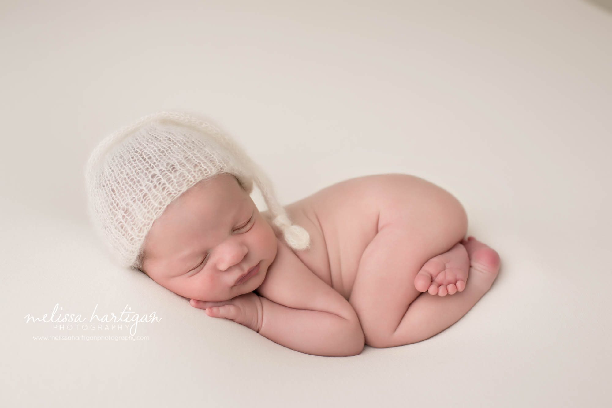 newborn baby boy posed on tummy on cream backdrop wearing knitted cream sleepy cap