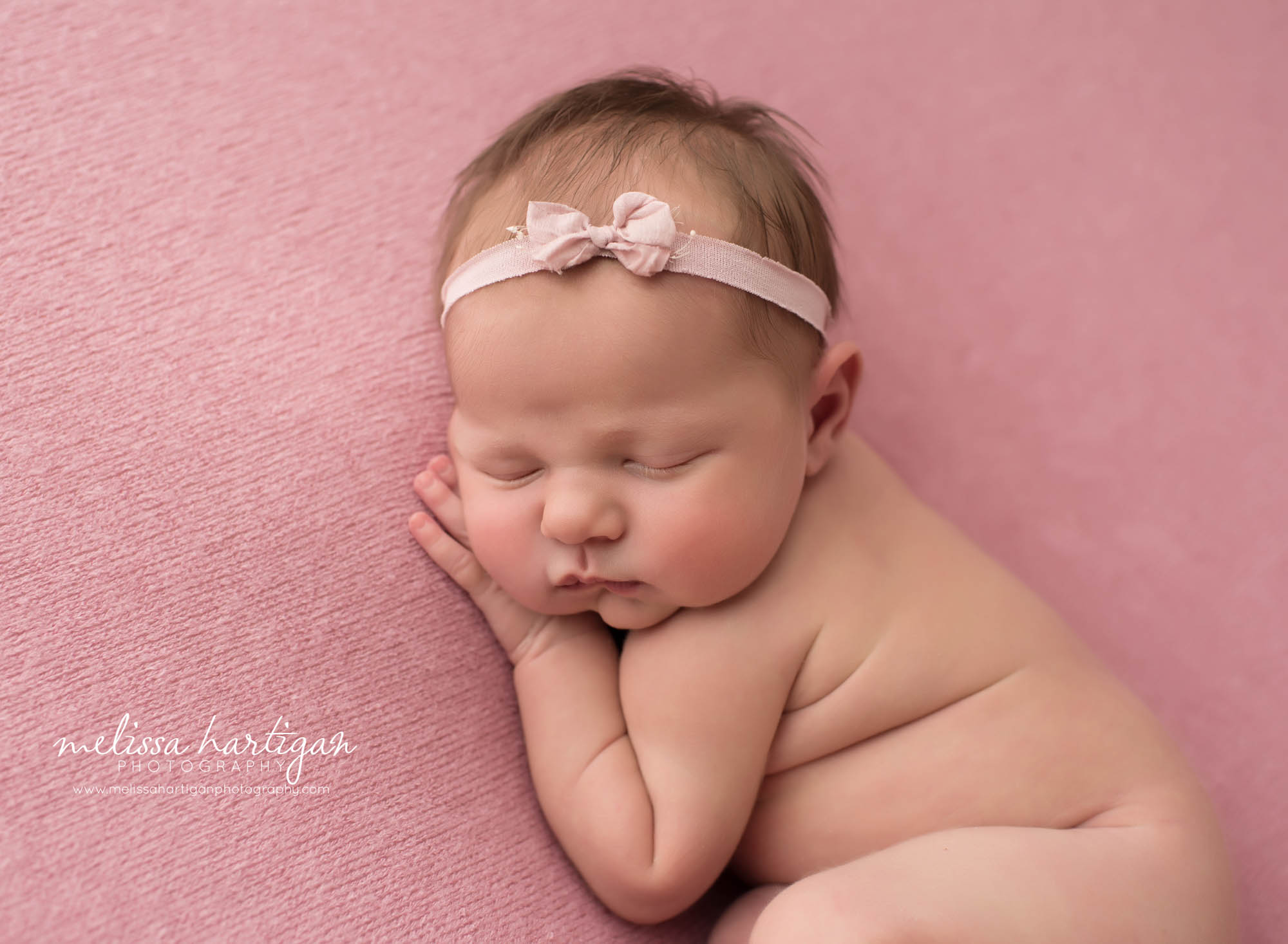 wborn baby girl posed on pink backdrop with hand under cheek wearing pink bow headband
