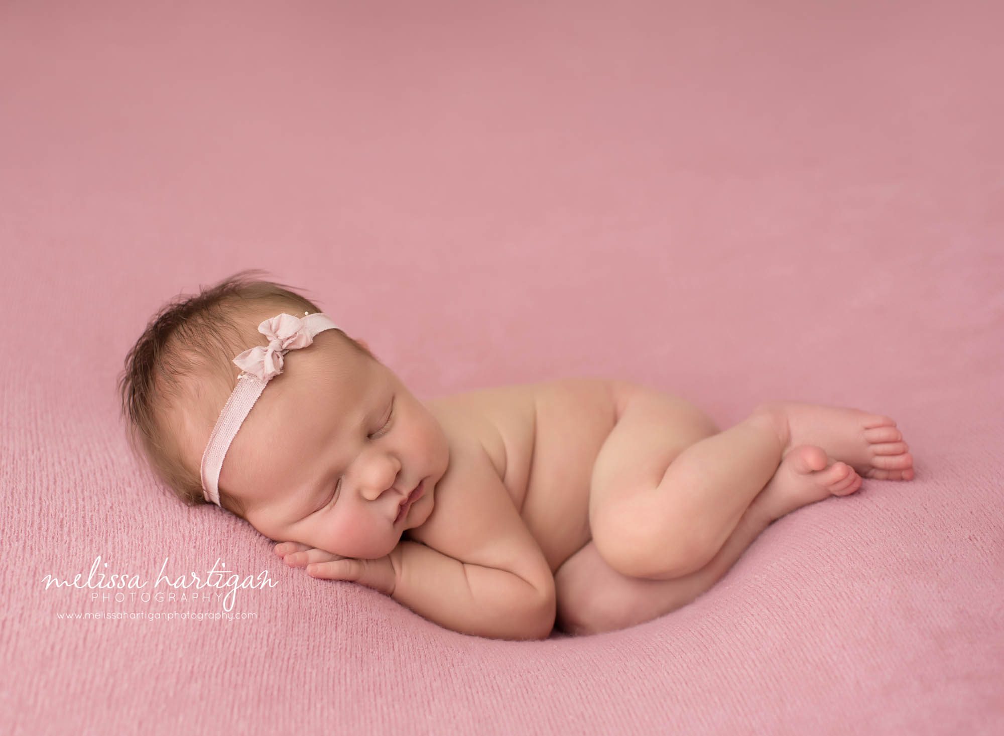 newborn baby girl posed on pink backdrop with hand under cheek wearing pink bow headband new london county baby photography