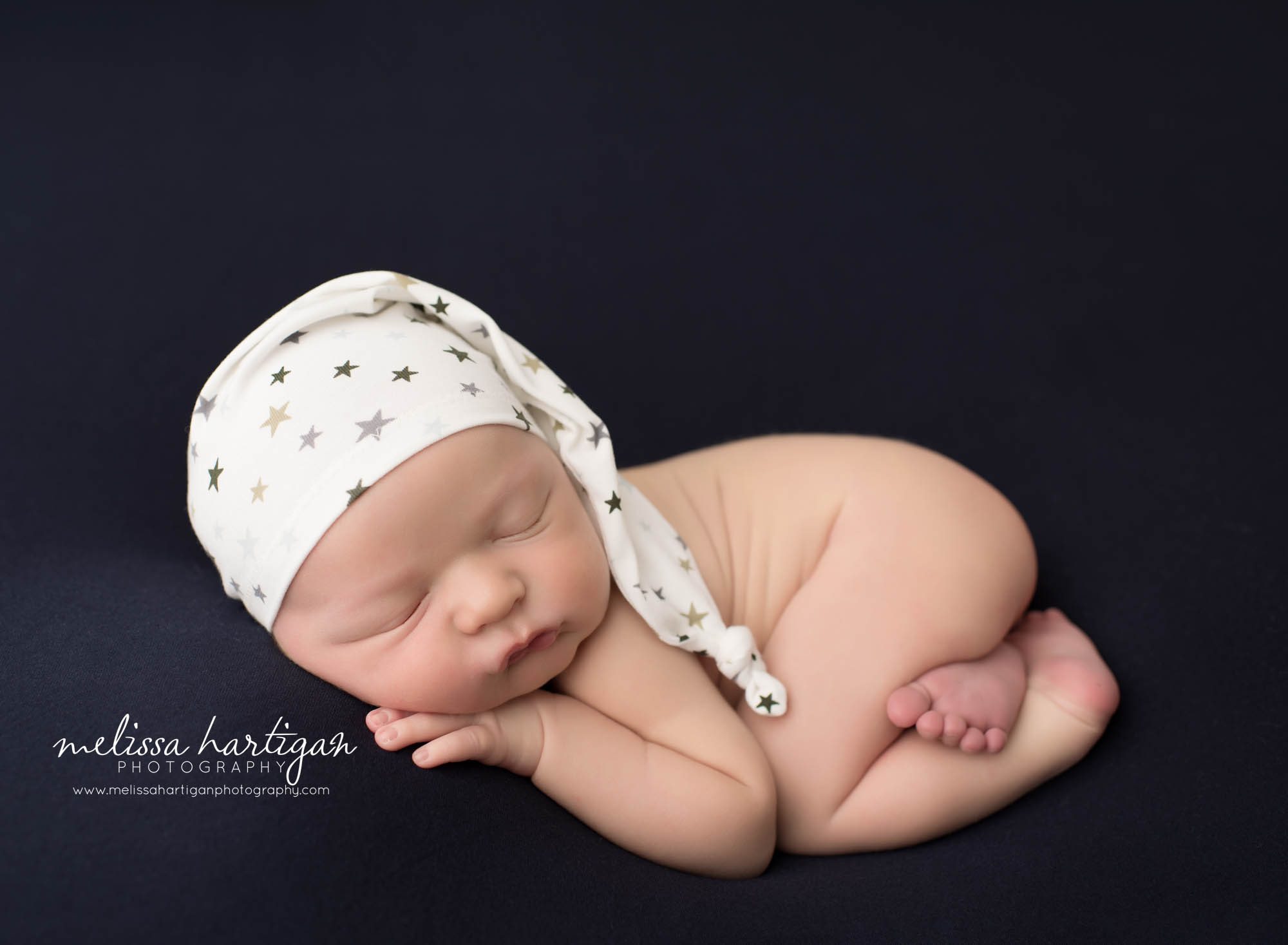 newborn baby boy posed on navy blue backdrop wearing white sleepy cap with stars