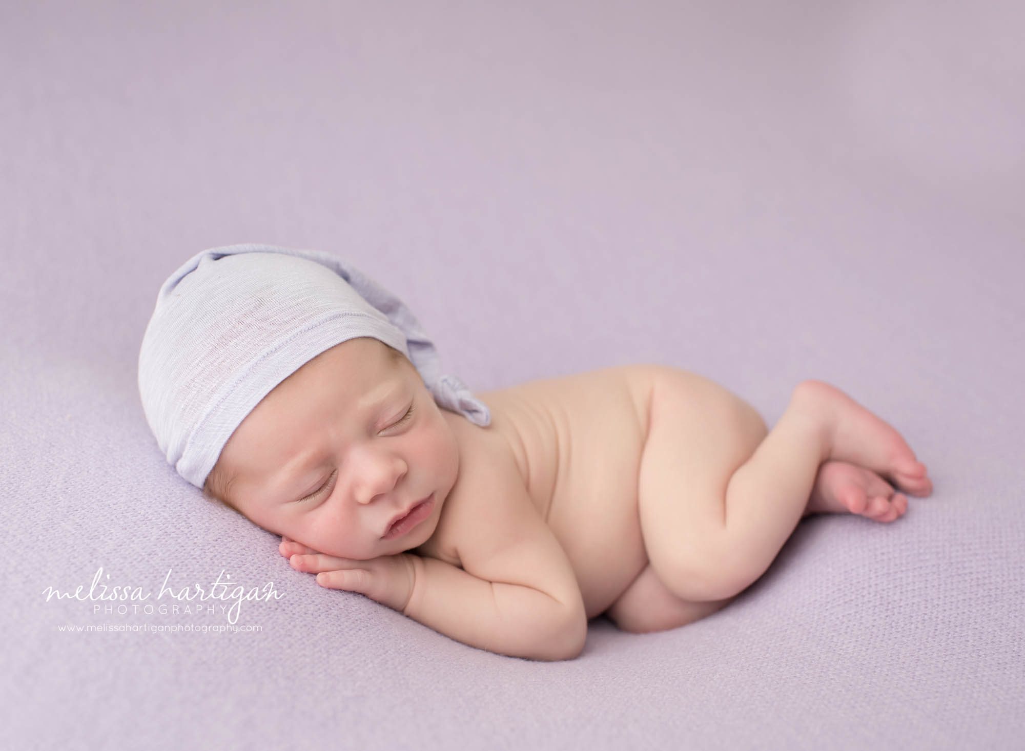 newborn baby girl posed on side with hand under cheek wearing light purple sleepy cap