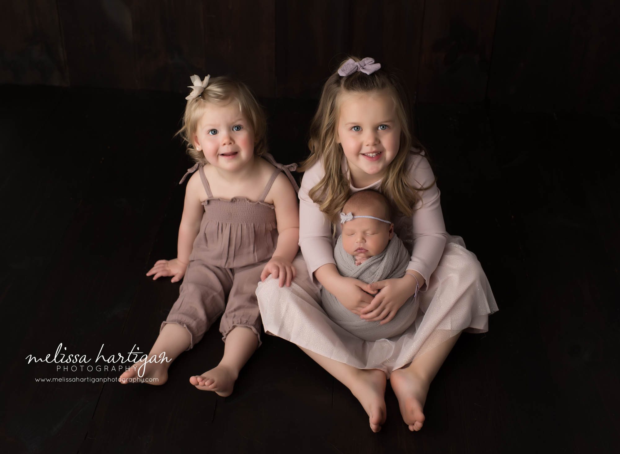 big sisters sitting on floor holding newborn sister swaddled in light green wrap family sibling pose rocky hill CT newborn photography