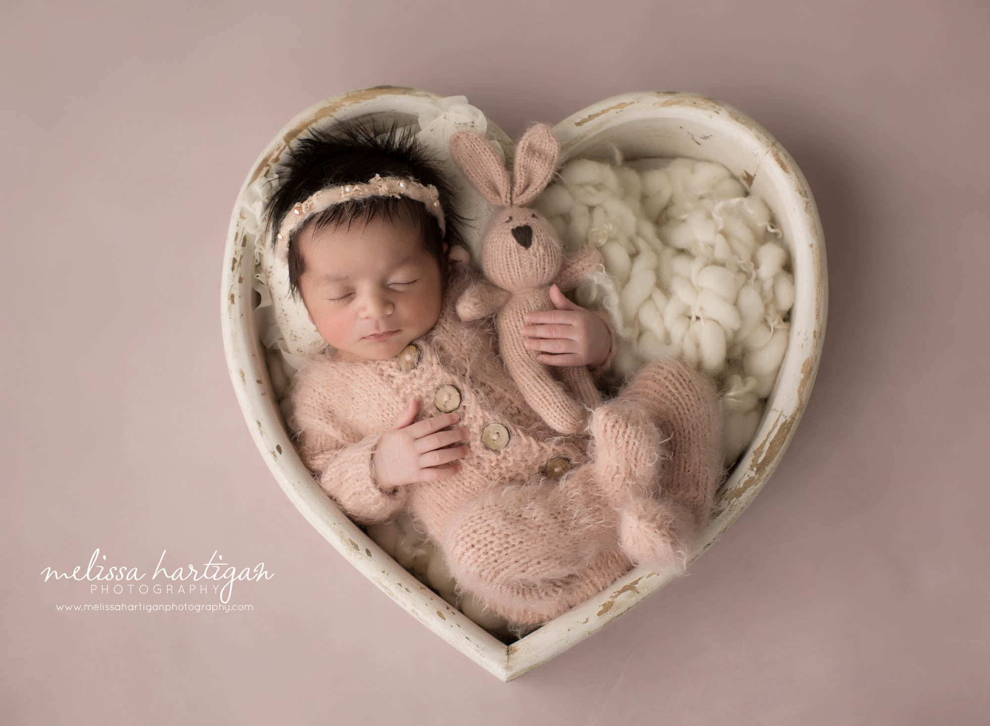 newborn baby girl wearing pink knitted outfit holding pink bunny prop wearing pink headband posed in cream wooden heart prop
