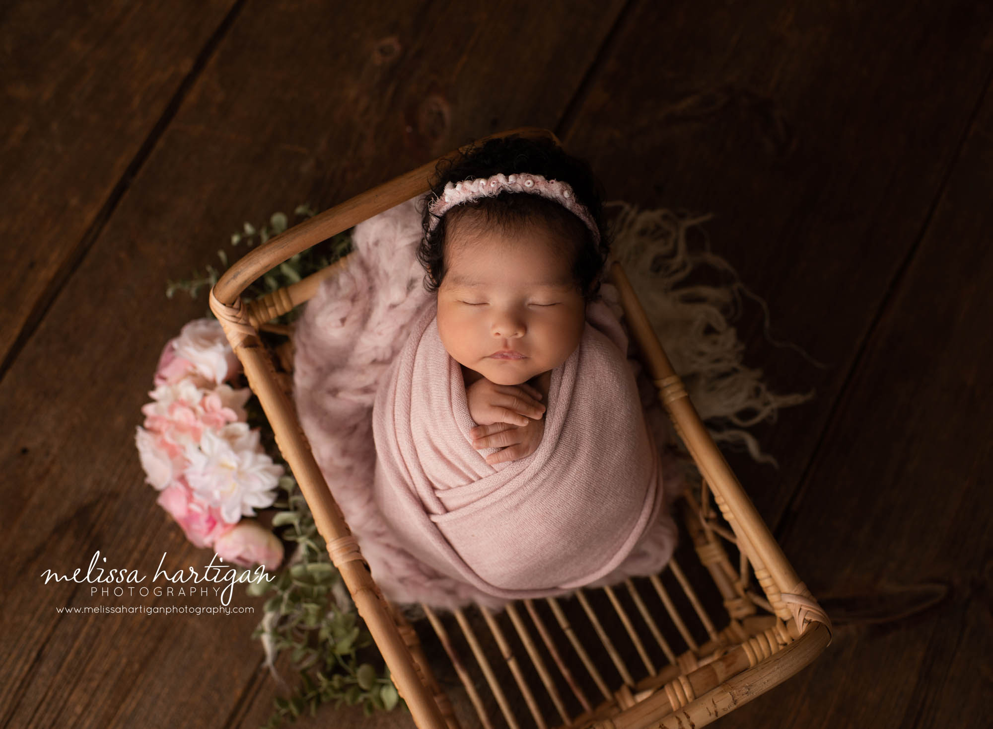 newborn baby girl wrapped in pink wrap posed in wicker basket with pink flowers