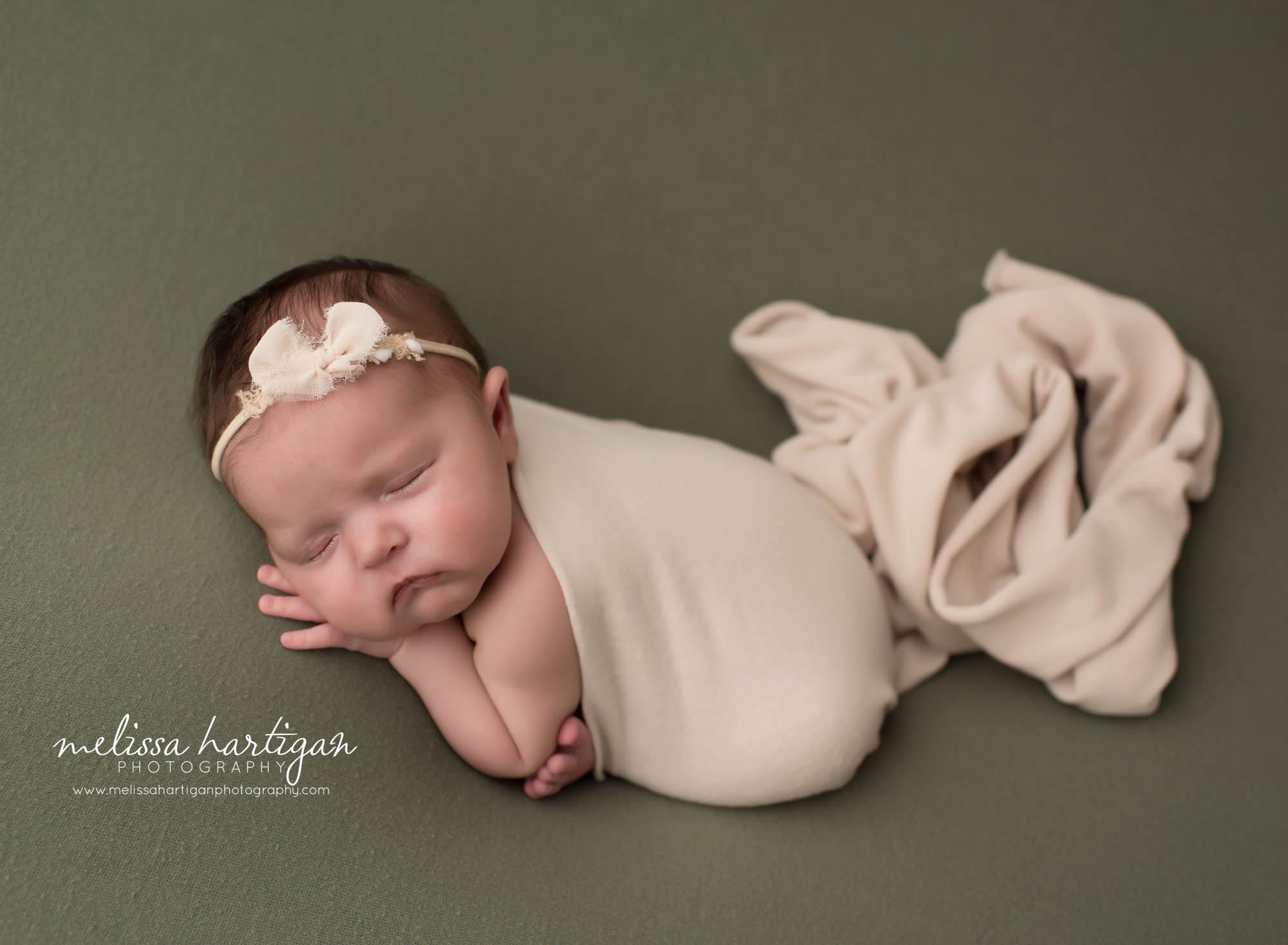newborn baby girl posed on side wearing cream bow headband and cream fabric draped and tucked under her sleeping peacefully