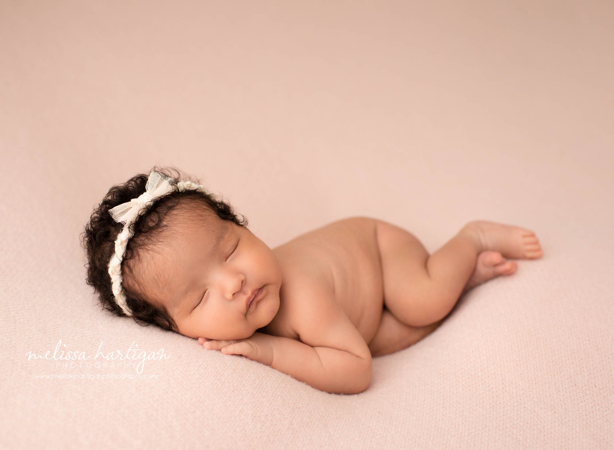 newborn baby girl posed on pink backdrop on side with hand under chin wearing cream bow headband newborn photography East Hartford