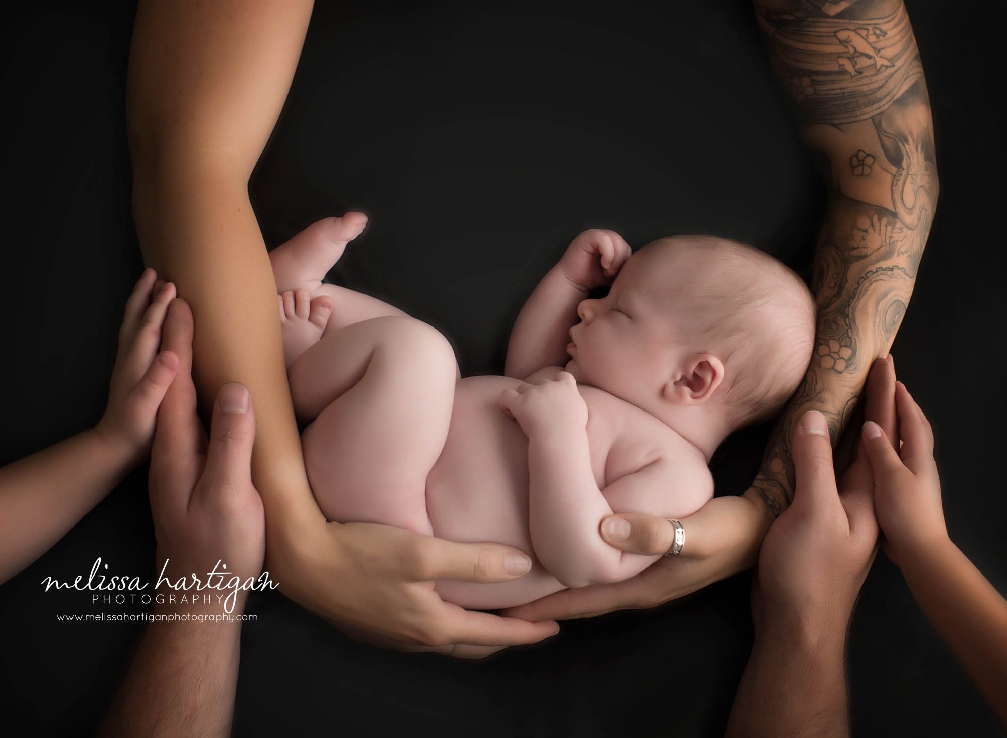 newborn baby boy posed on black backdrop with moms arms cradling him and dads hands and two big sisters hands holding him