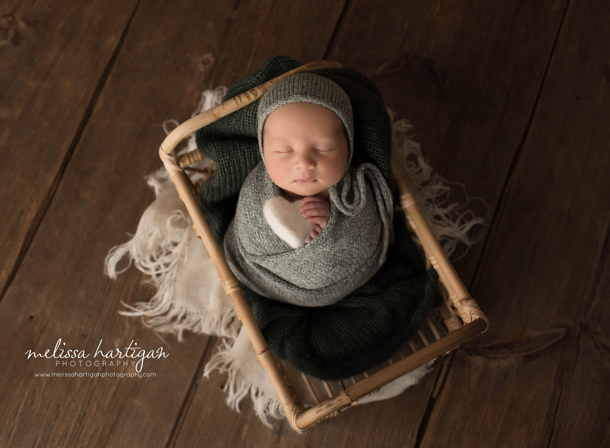 newborn baby boy posed in basket wearing knitted bonnet and wrapped in matching colored wrap holding felted heart prop