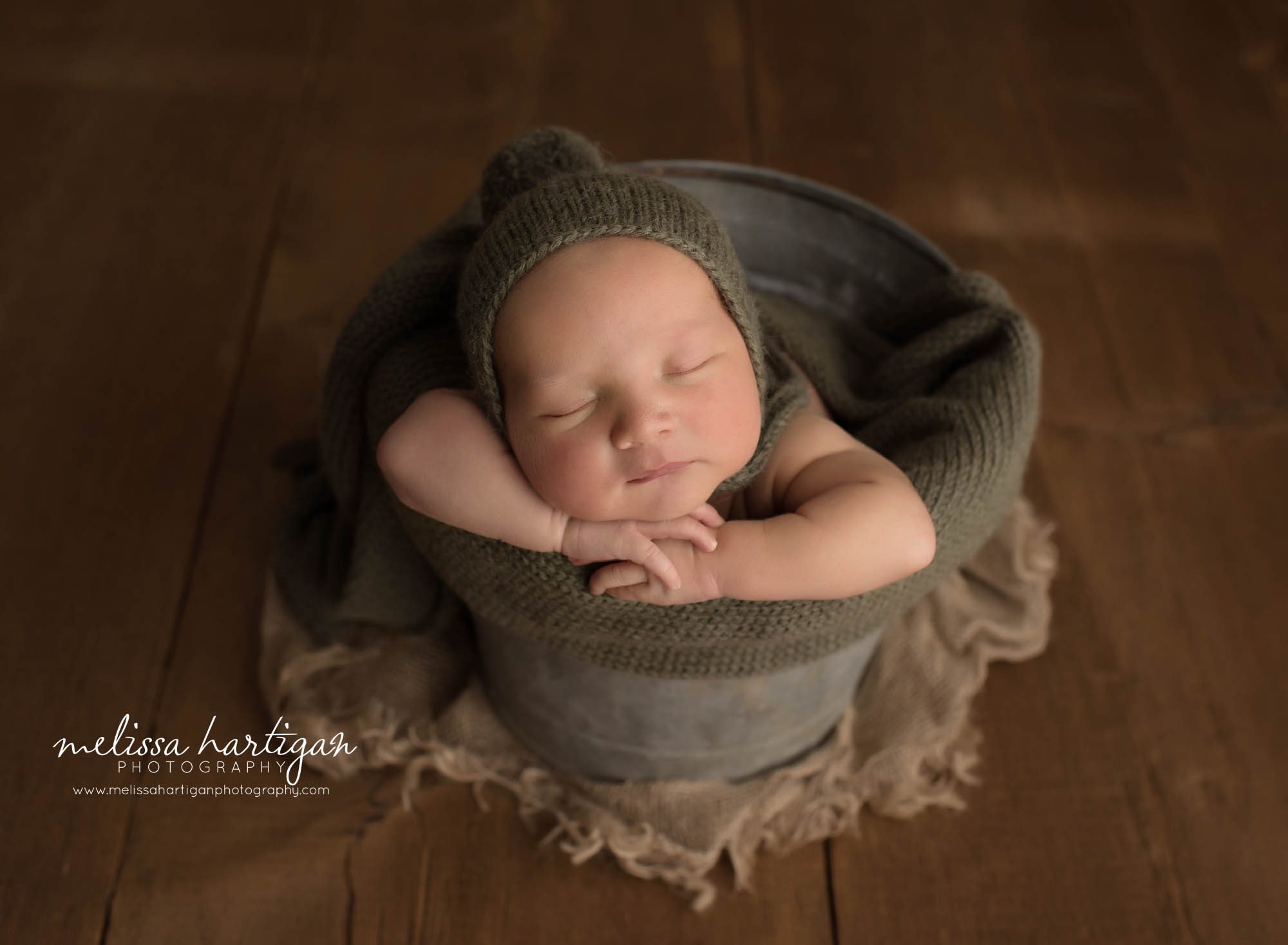 newborn baby boy posed in metal bucket wearing green pom sleepy cap bonnet