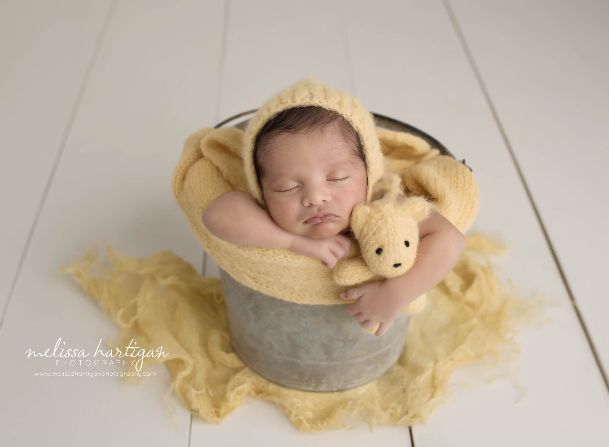 newborn baby boy posed in metal bucket with yellow knitted wrap wearing yellow knitted bonnet with matching yellow teddy bear prop