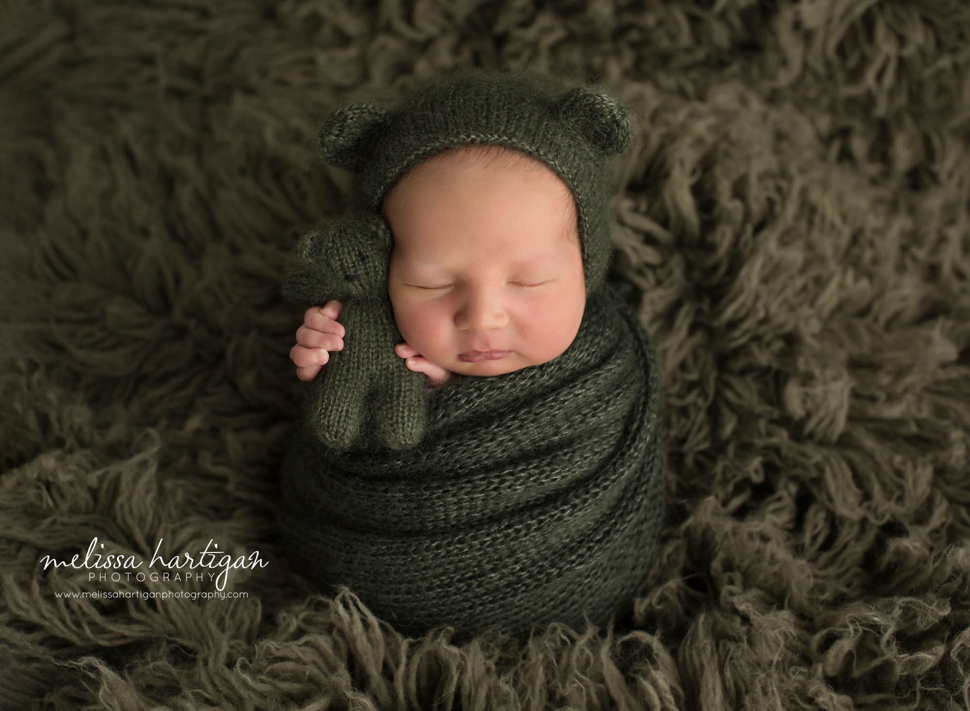 newborn baby boy wrappe din dark green knitted wrap posed on green flokati wearing knitted bear bonnet holding matching knitted teddy bear Newborn photography CT
