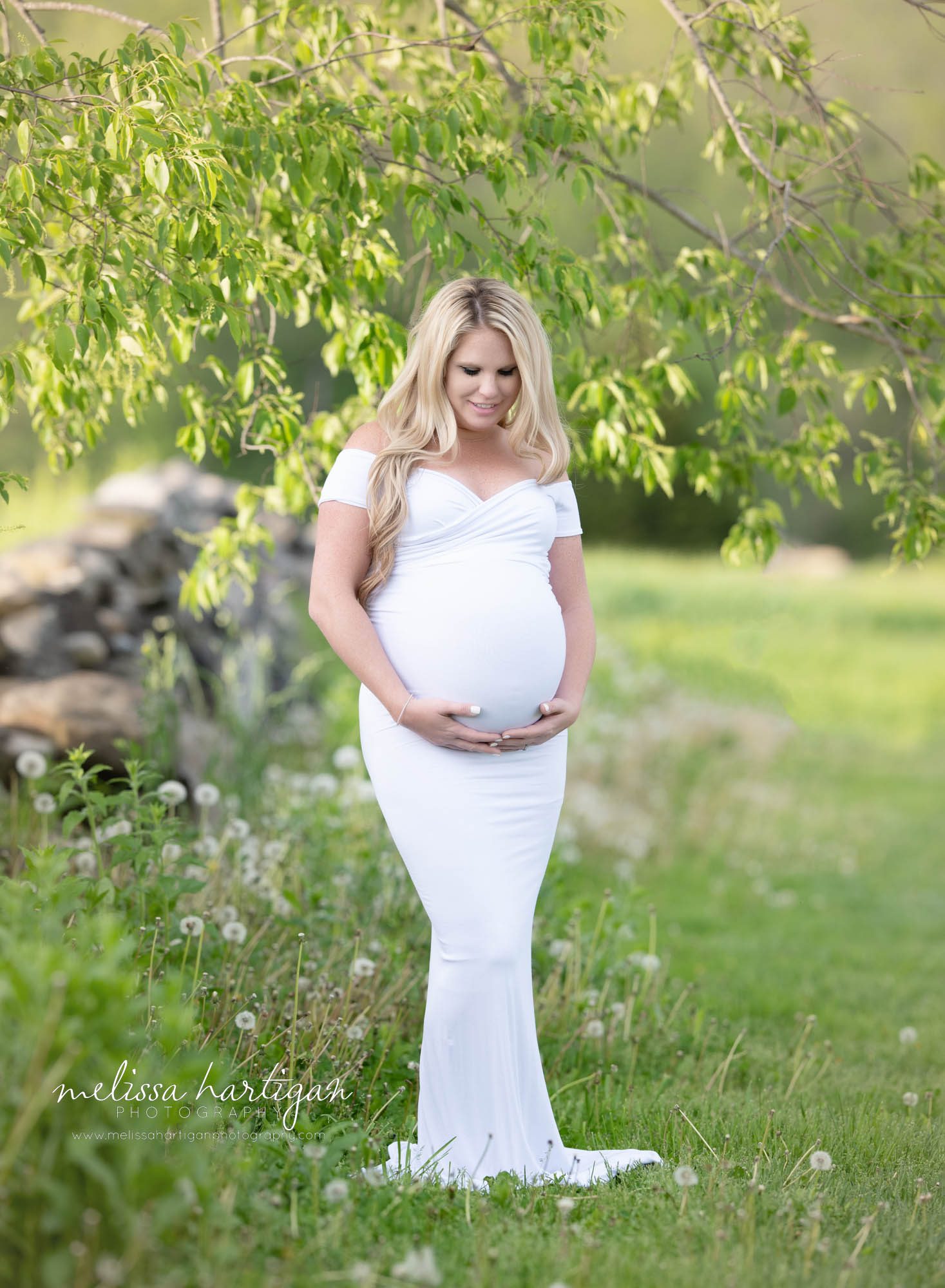 pregnant mom holding and looking down at baby bump wearing long white form fitted sweetheart neckline maternity gown