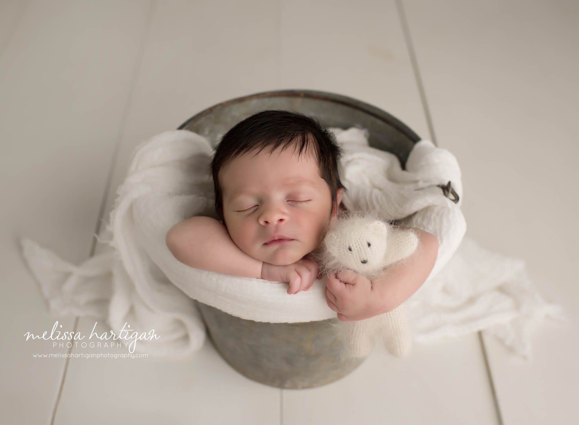 newborn baby boy posed in metal bucket with white draping layer and white knitted teddy bear