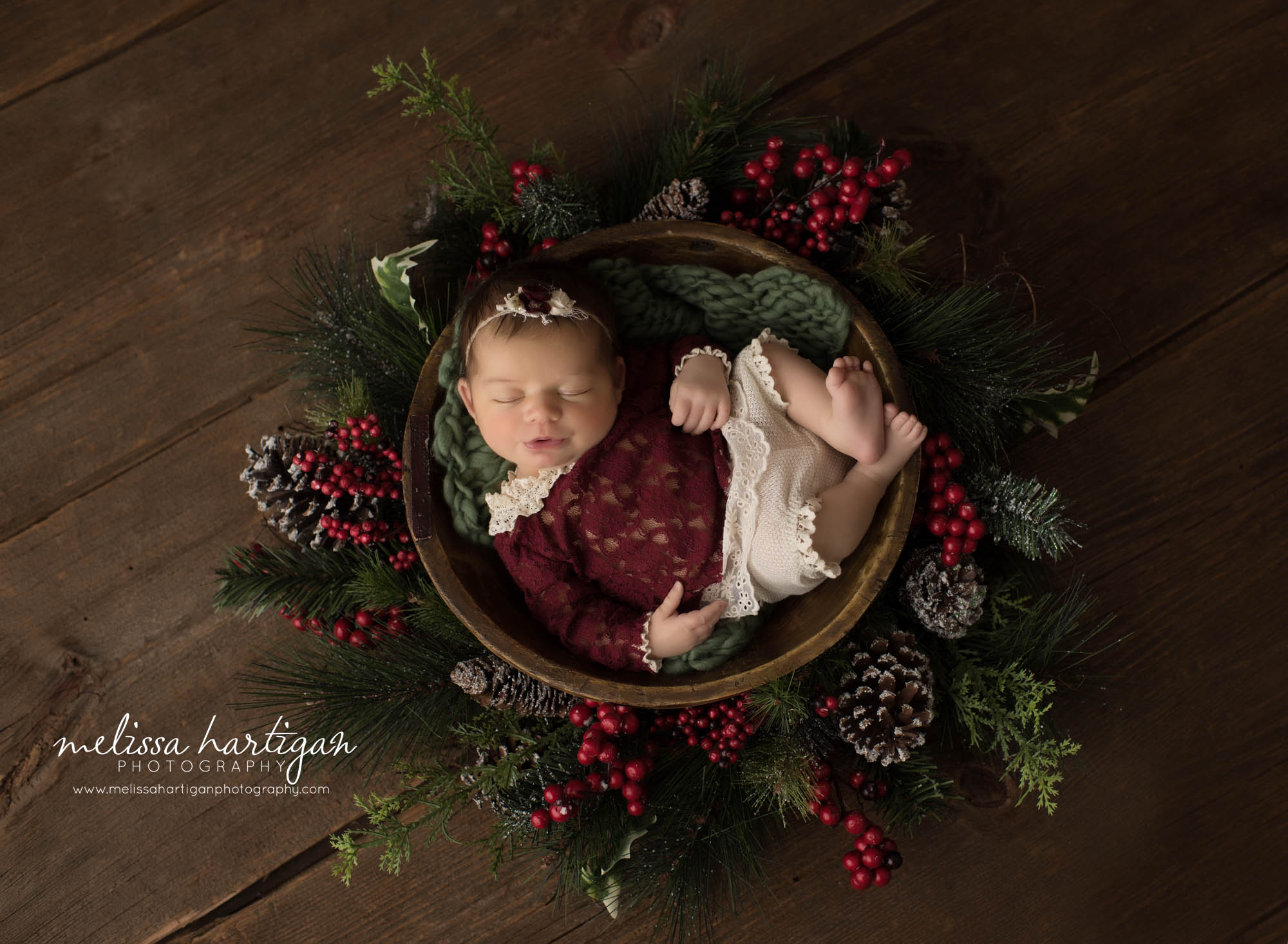 newborn baby girl posed in wooden bowl wearing red baby girl outfit posed with christmas garland and pinecones