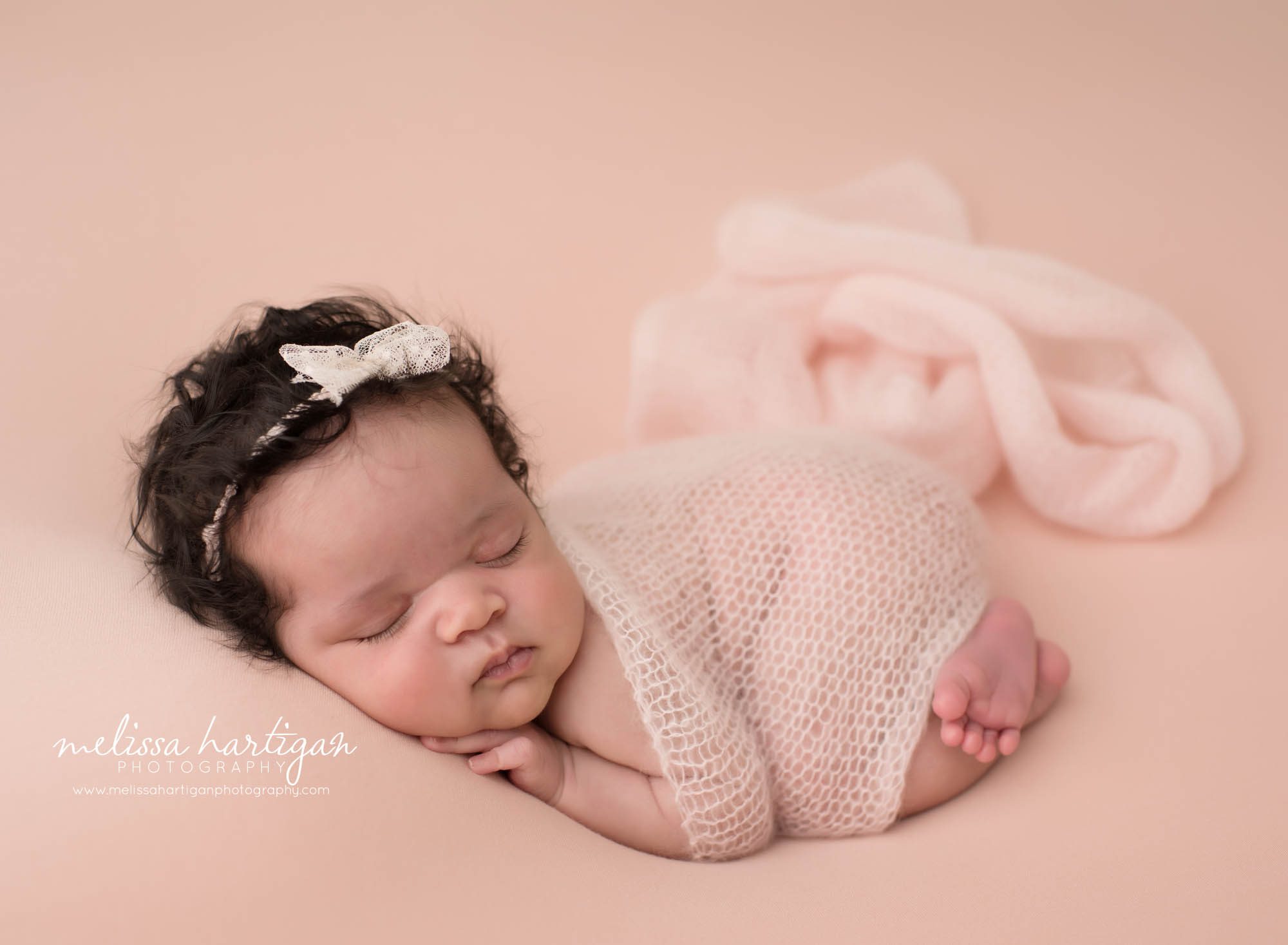 newborn baby girl posed on tummy wearing bow headband and pink knit wrap draped over