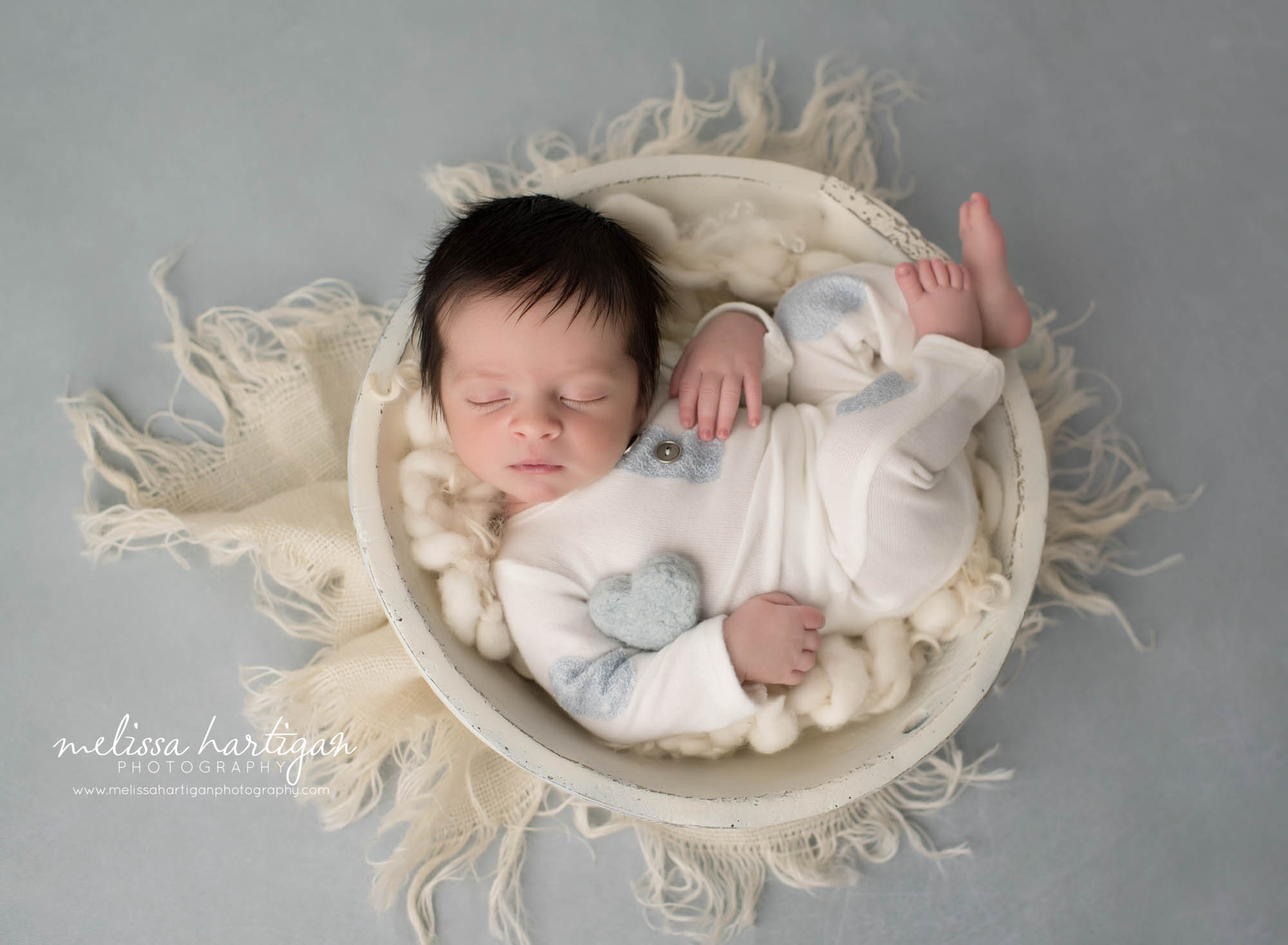 newborn baby boy posed in cream wooden bowl wearing white newborn boy outfit with gray felted heart prop