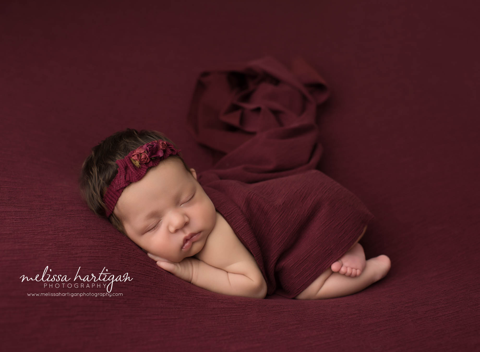 Newborn baby girl posed on burgundy backdrop wearing headband posed on tummy tolland newborn photography