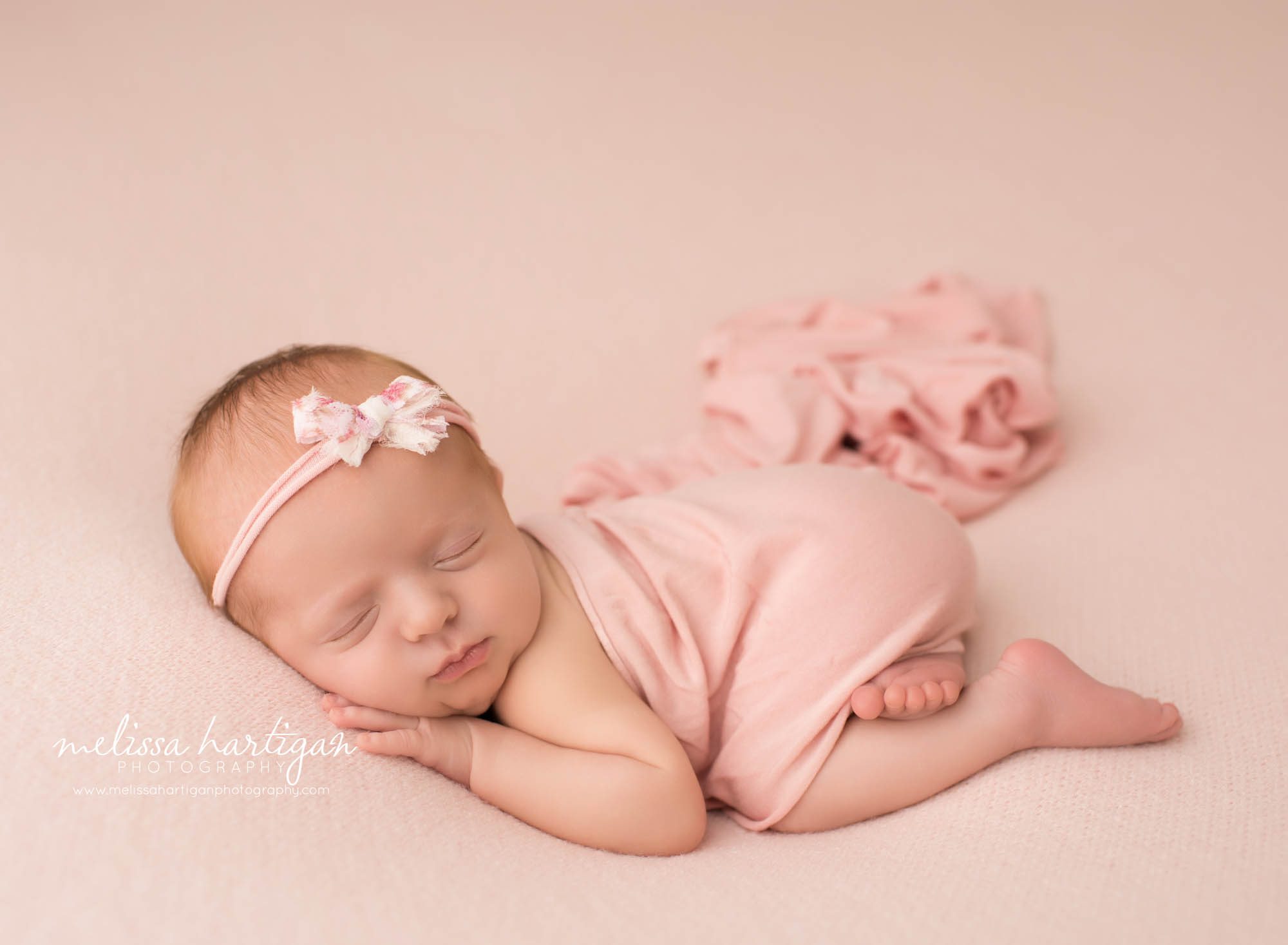 newborn baby girl posed on tummy sleeping wearing pink and white bow headband pink wrap draped over her back