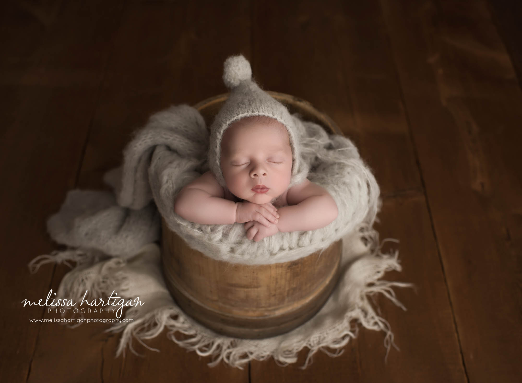 newborn baby boy posed in wooden barrel with gray knited wrap layer wearing pom pom bonnet