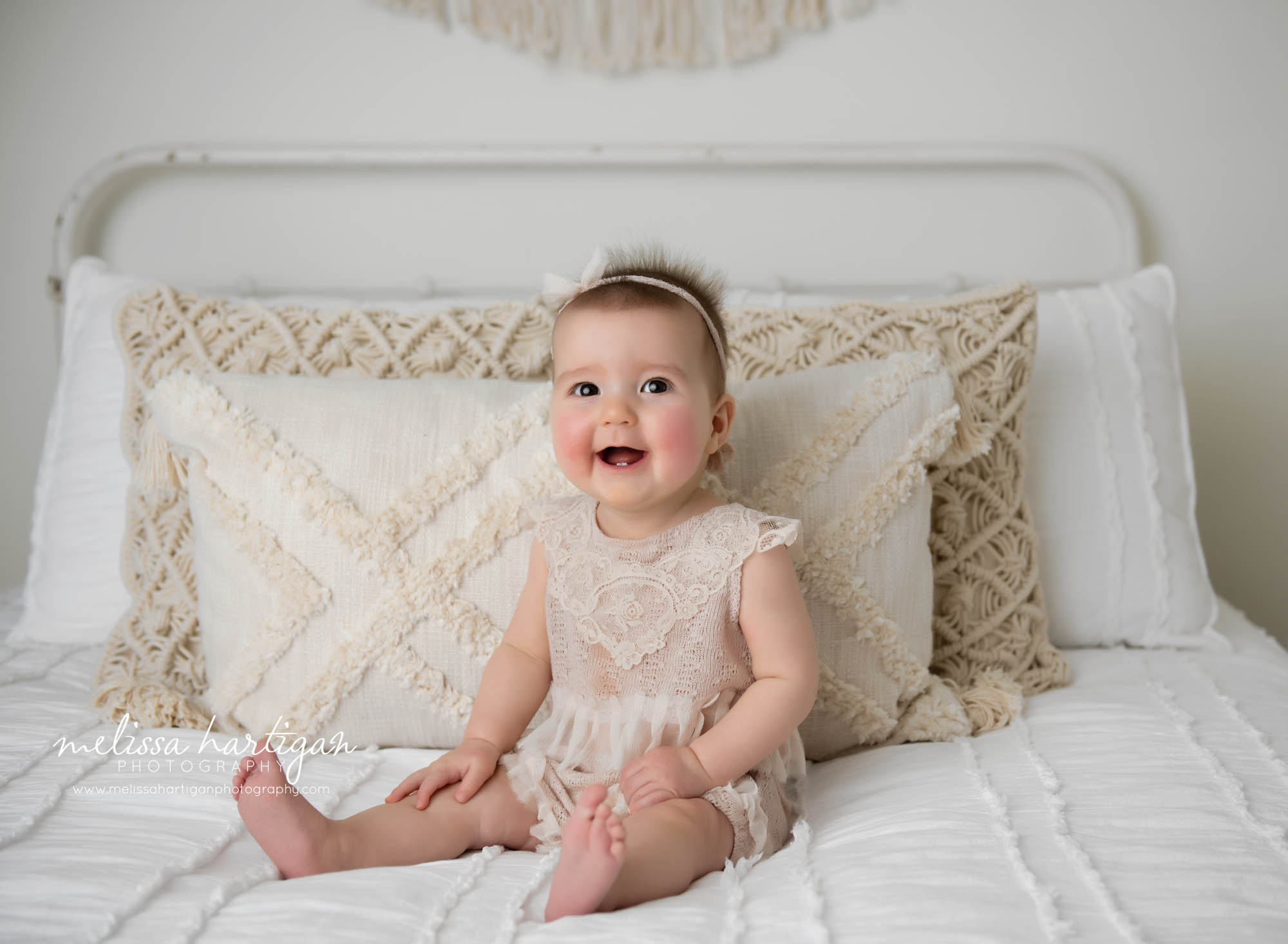 baby girl sitting up on bed smiling showing her two bottom teeth