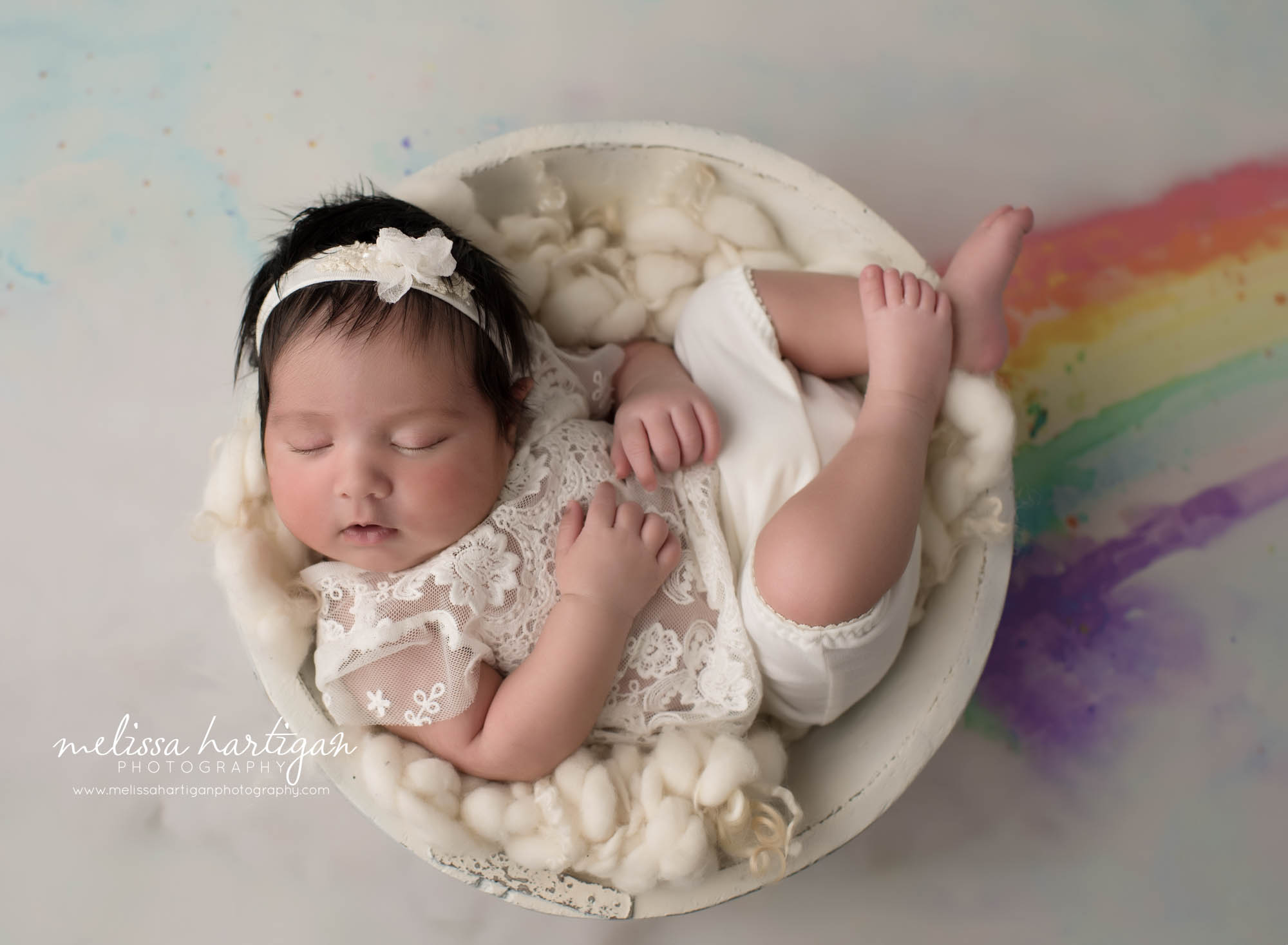 newborn baby girl posed in wooden bowl wearing cream outfit with rainbow coming out side