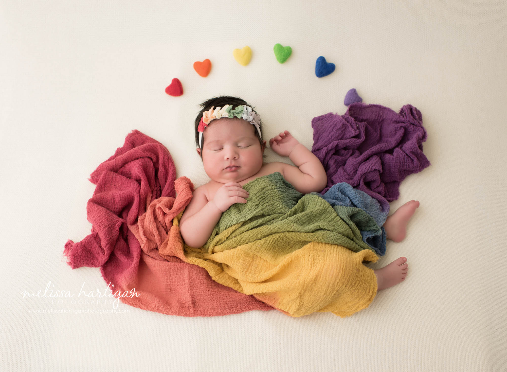 newborn baby girl posed on cream backdrop with rainbow colored wrap and rainbow felted hearts