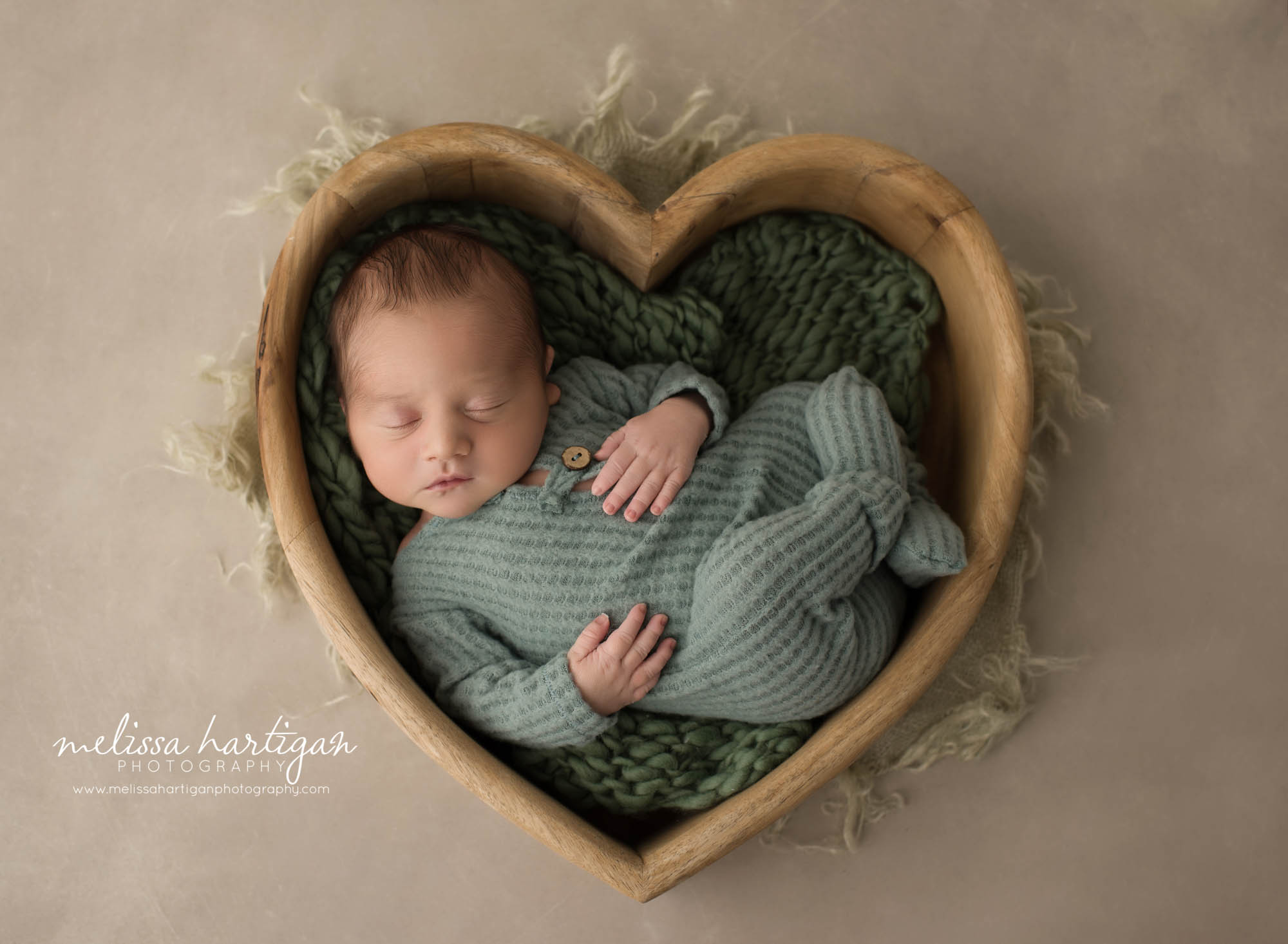 newborn baby boy posed in wooden heart shaped prop wearing green footed sleeper