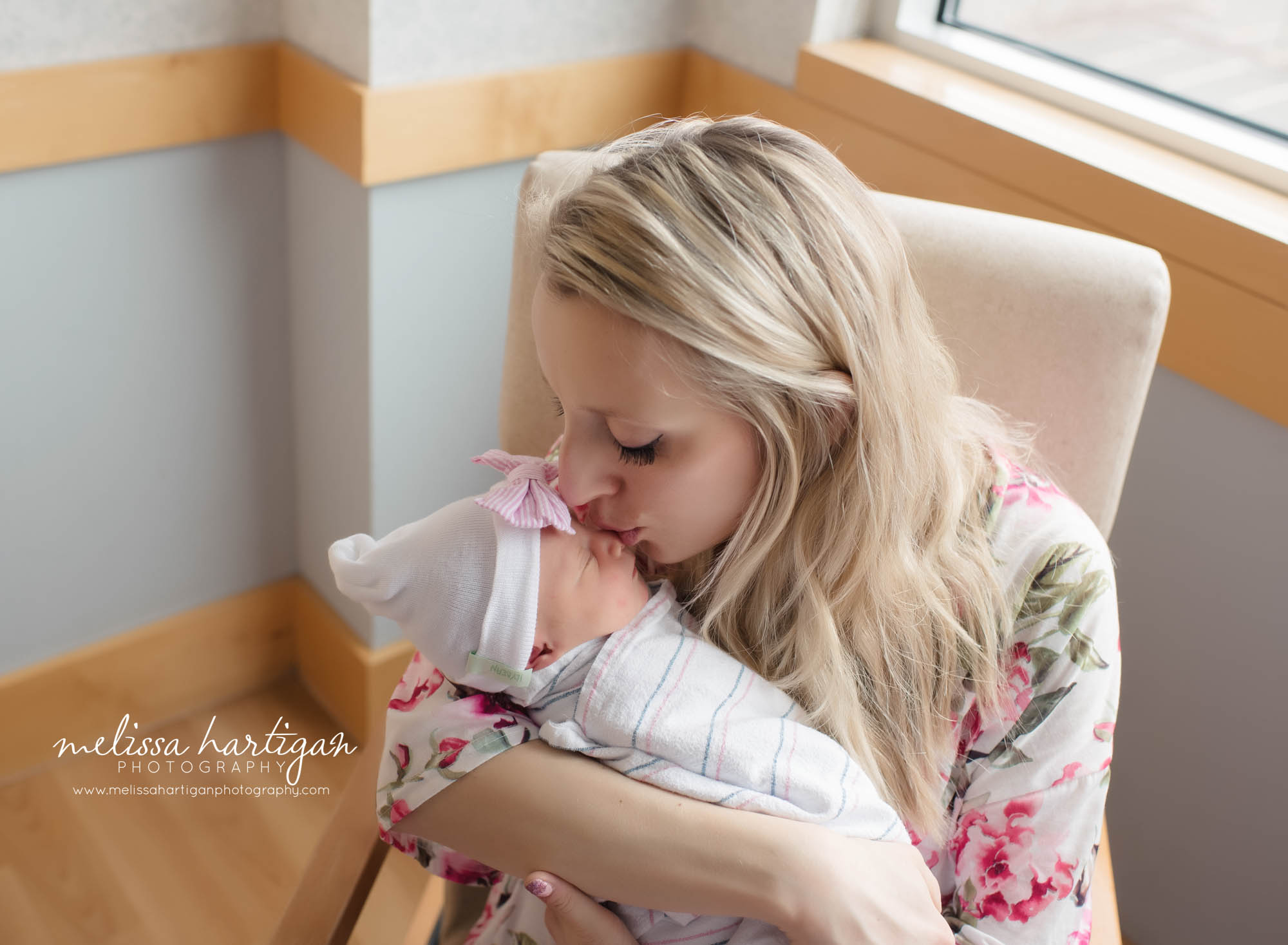 mom holding newborn daughter sitting in hospital chair giving kiss to newborn daughter