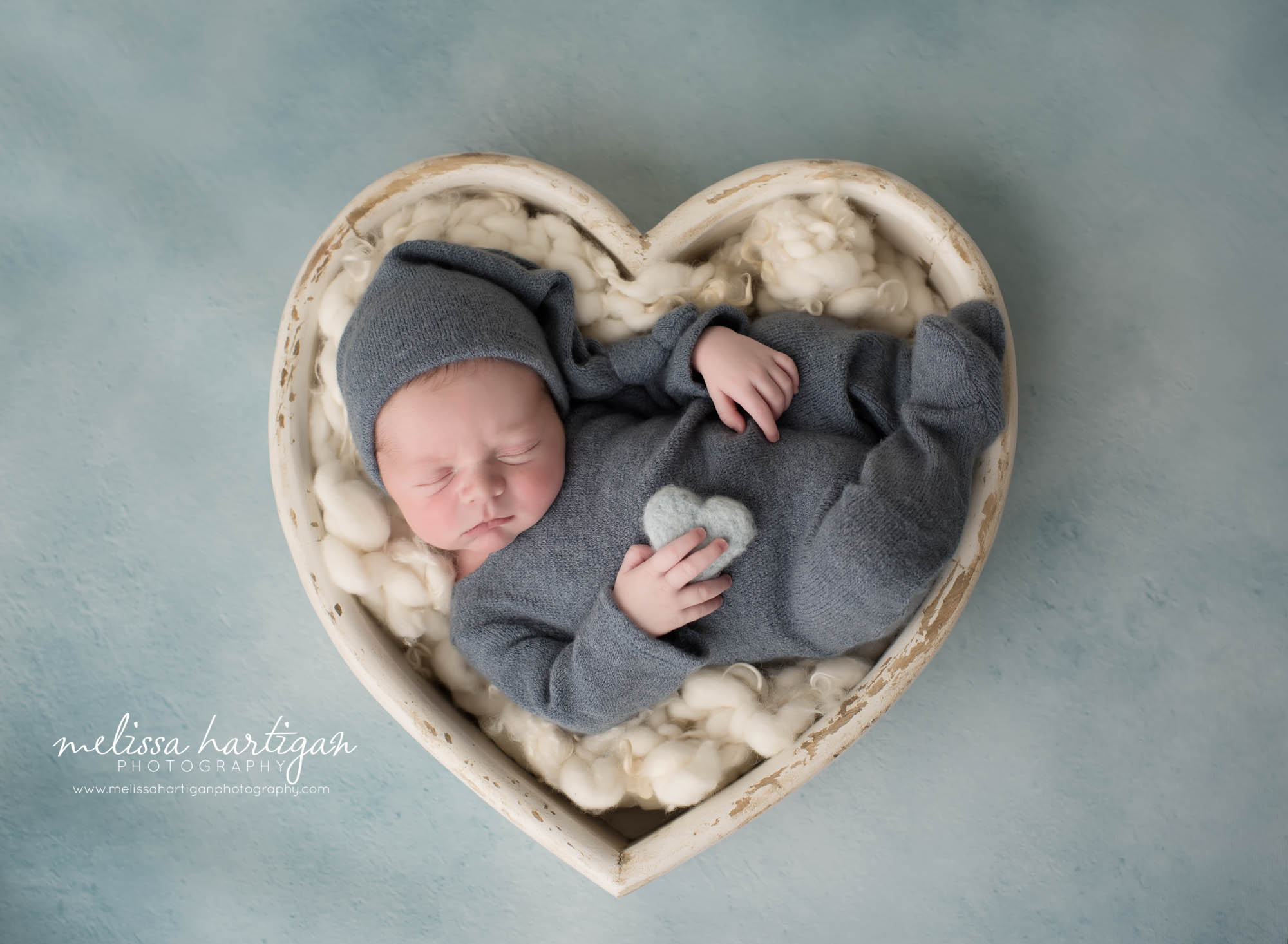 newborn baby boy posed in cream wooden heart prop wearing blue gray outfit and sleepy cap